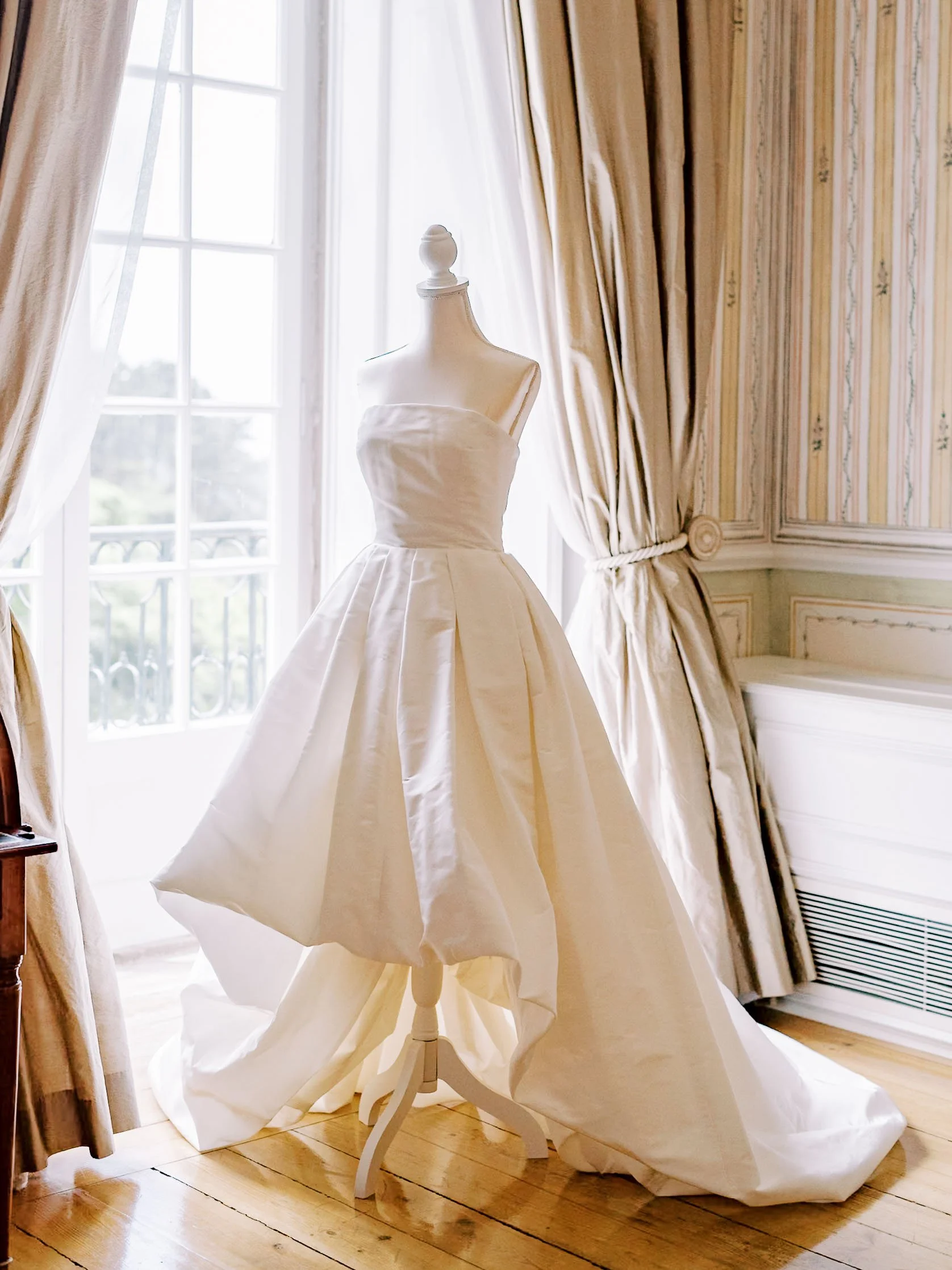 Elegant ivory wedding dress displayed on a mannequin in a bright room with large windows, floral wallpaper, and beige curtains.