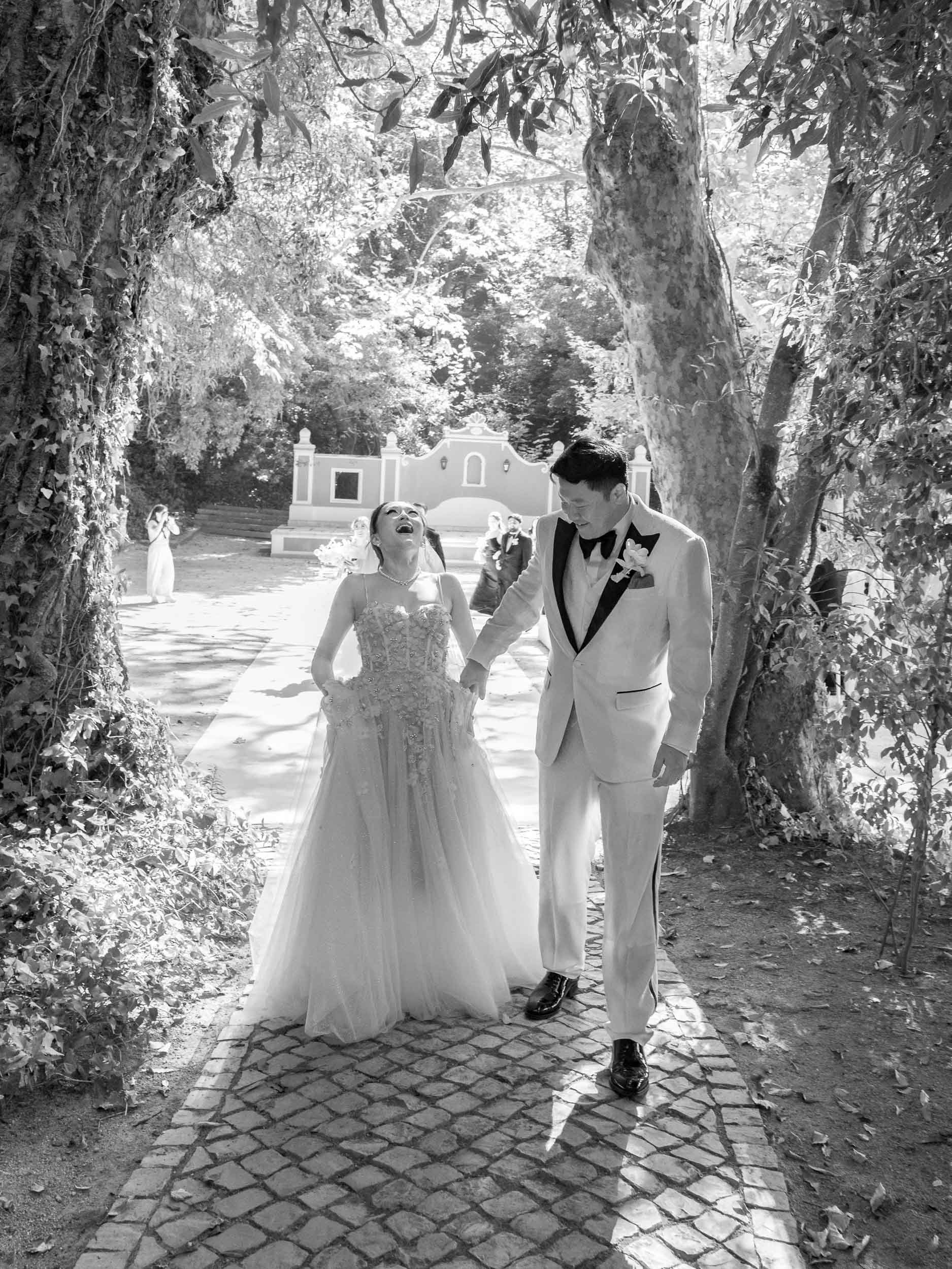 Black and white photo of a bride and groom holding hands and walking outdoors under trees, smiling and looking at each other, with wedding guests in the background at Quinta da Bella Vista