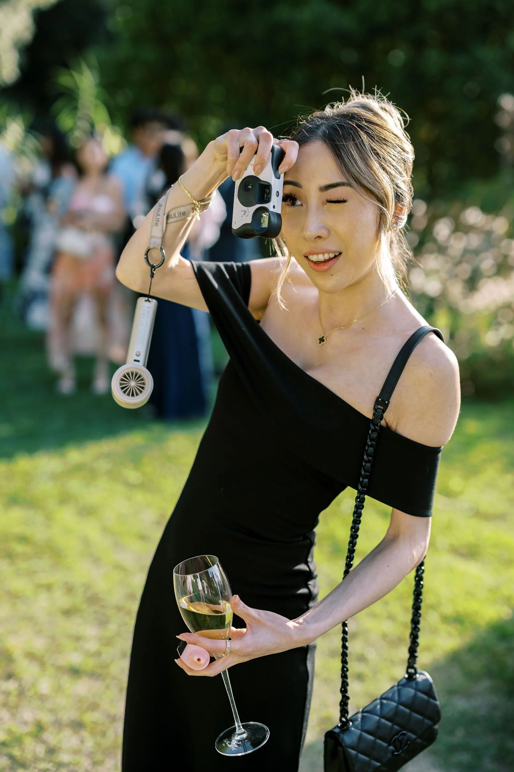 A bridesmaid in a black off-shoulder dress taking a photo with a camera at cocktail hour. She is holding a glass of white wine and a small fan, with a group of people in the background on a sunny day at Quinta da Bella Vista.