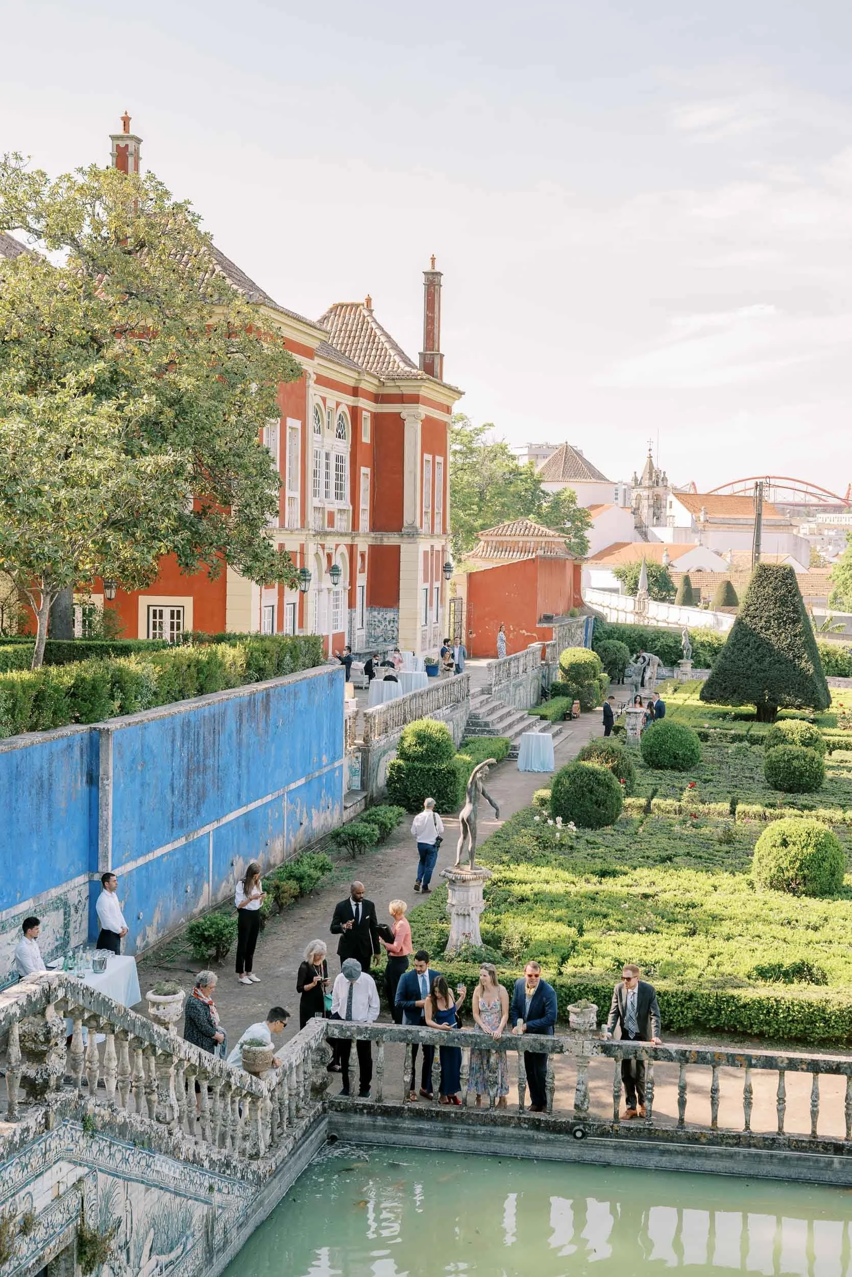 A garden party with guests mingling near a fountain, tall trimmed hedges, a grand red-orange building, and statues, under a bright sky.