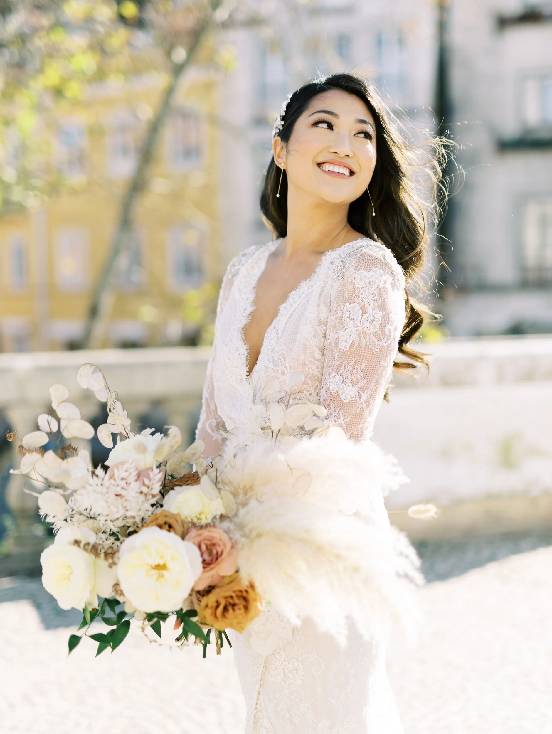 A bride in a white lace wedding dress holding a bouquet of white, yellow, and pink flowers, standing outdoors with trees and buildings in the background.