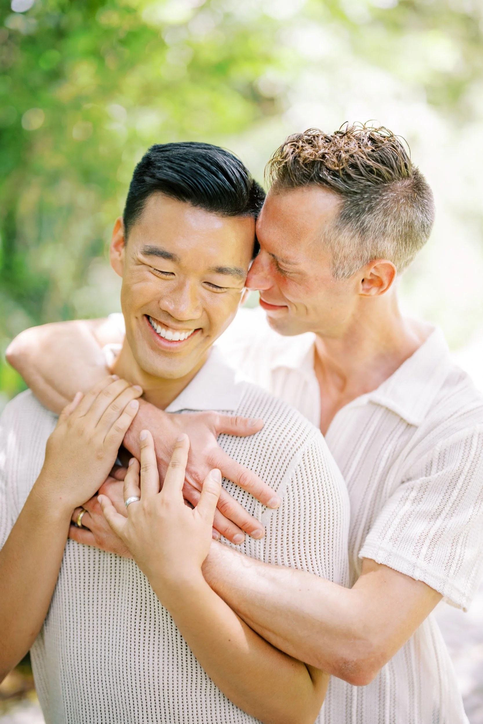 Two grooms smiling and embracing outdoors with green foliage in the background.