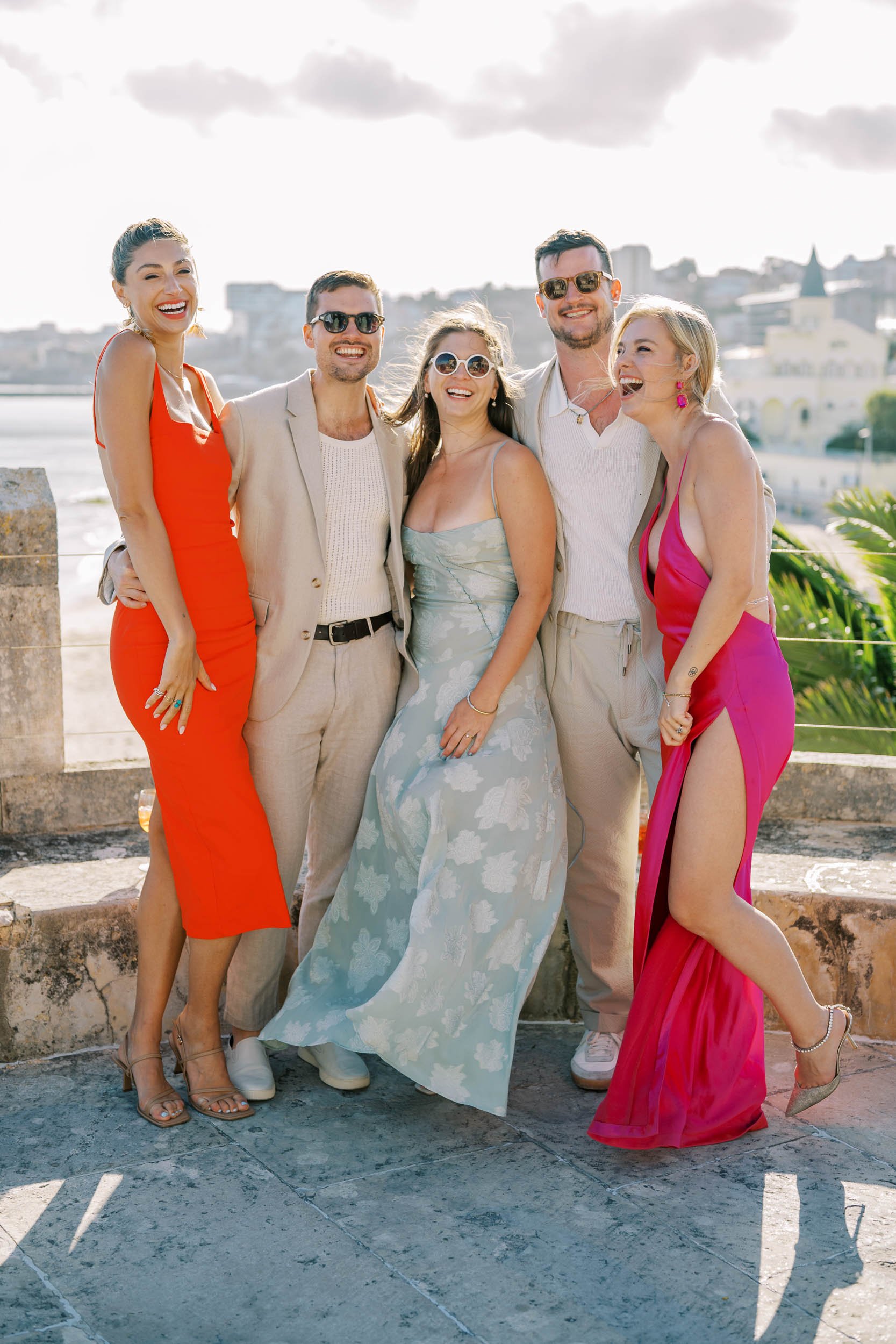 Group of five wedding guests dressed in stylish summery outfits, smiling and posing outdoors against a scenic cityscape background.