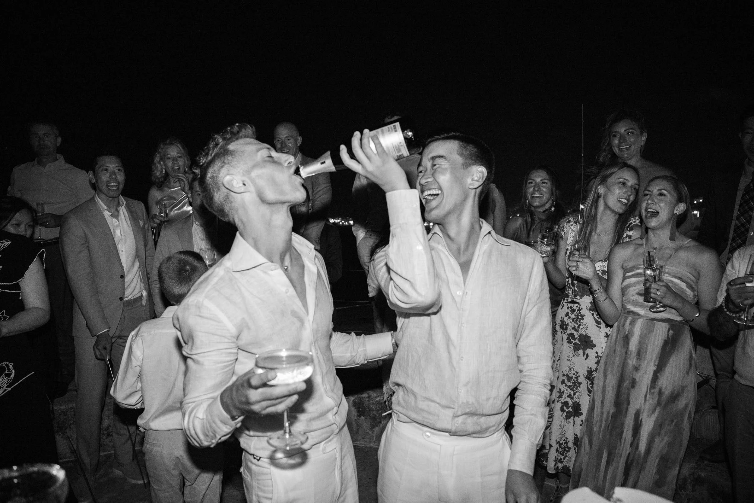 Two Grooms at a party, one pouring a drink into the other's mouth while surrounded by smiling people holding drinks, all dressed in formal or semi-formal attire at night.