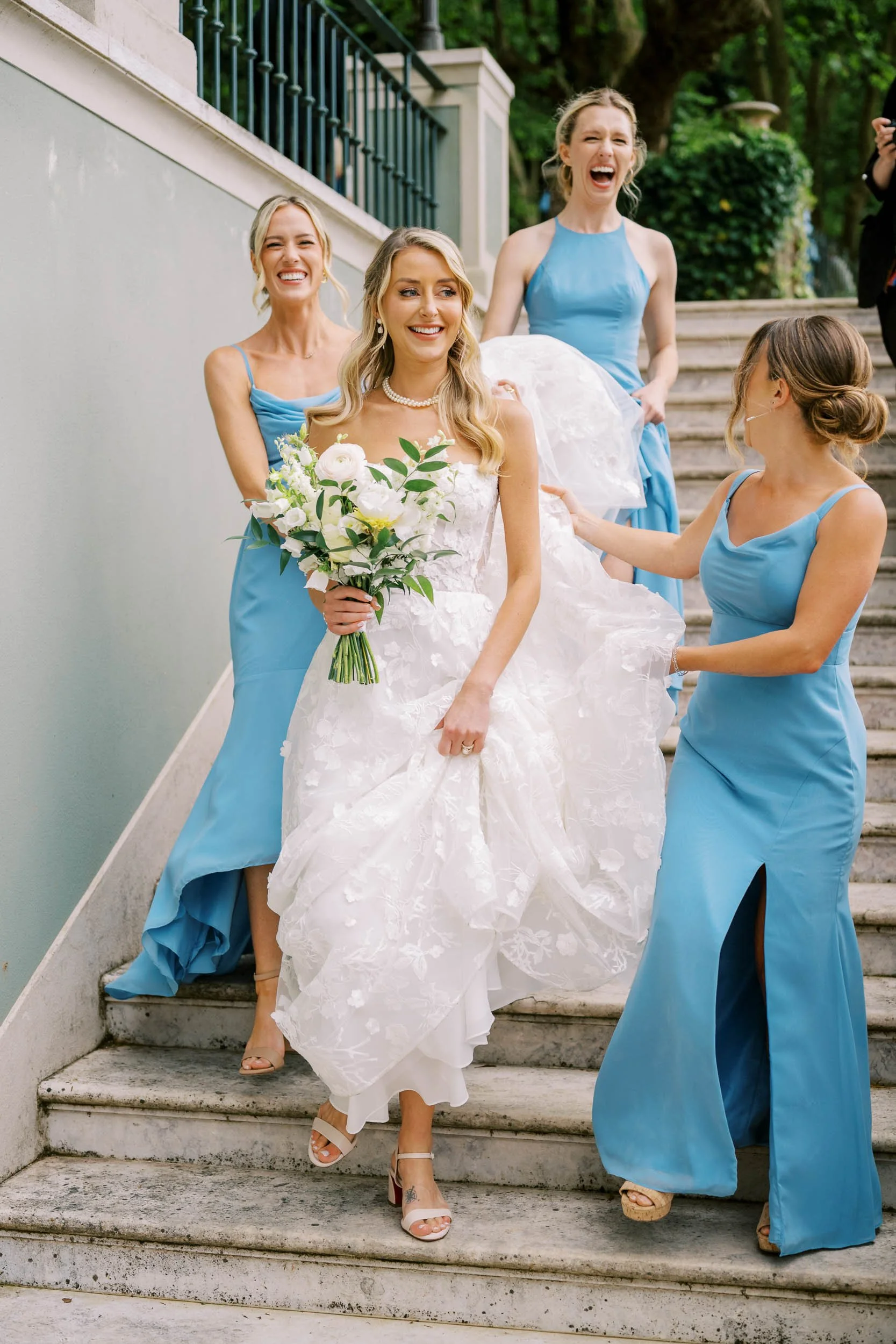A bride in a white wedding dress holding a bouquet, smiling as her bridesmaids in blue dresses assist her down the stairs, all laughing and enjoying the moment outside.