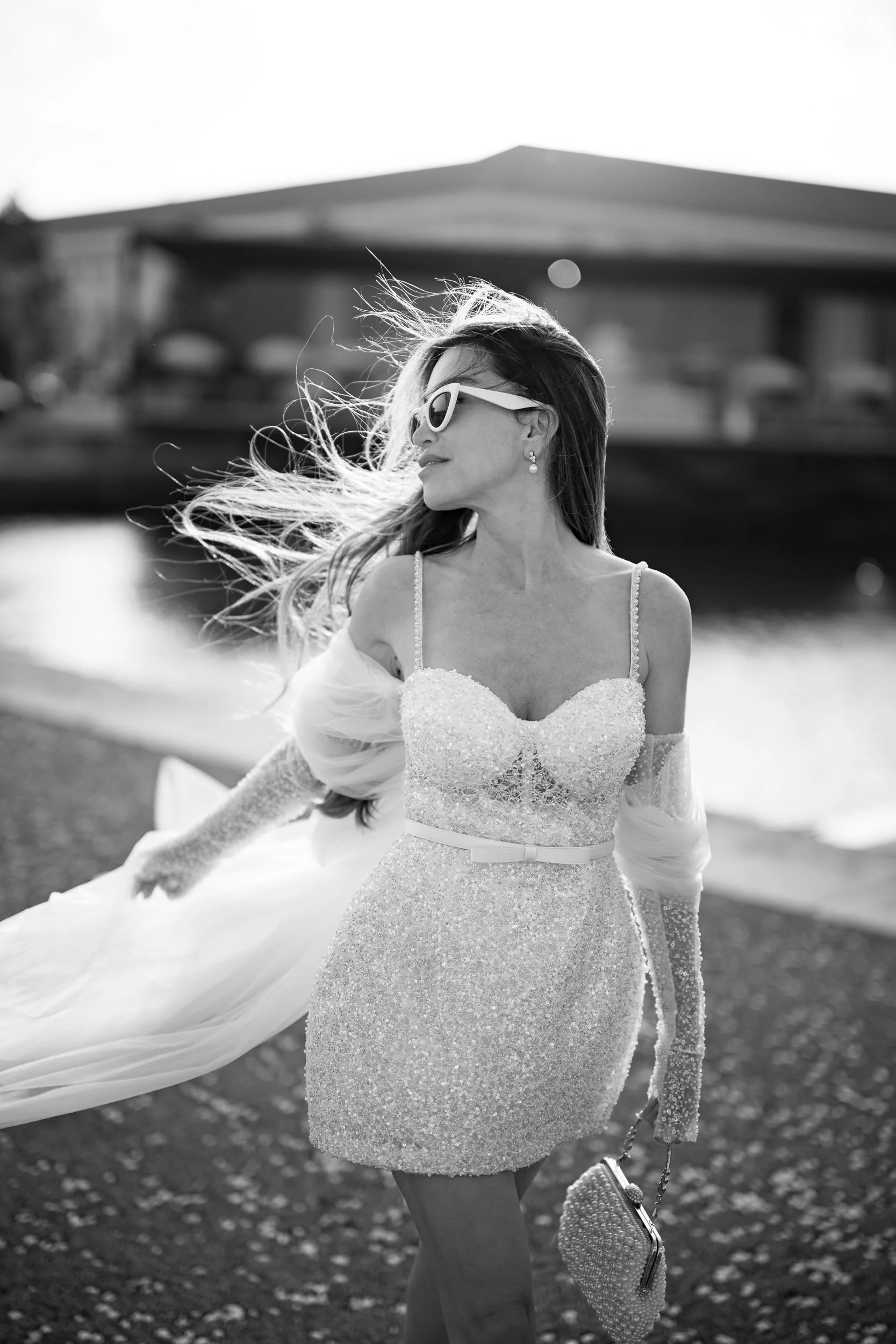 A bride wearing sunglasses and a sparkly, sleeveless dress with a tulle train, walking along a waterfront with her hair blowing in the wind.