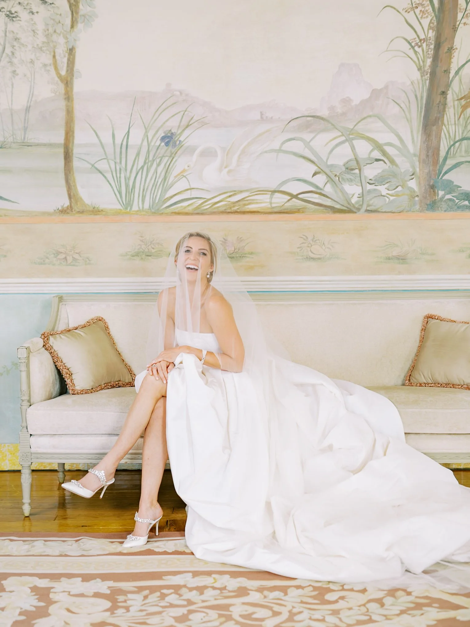 A bride sitting on a vintage sofa, wearing a white wedding dress and veil, smiling and laughing. The setting is indoors with a painted mural of nature scene behind her. at Palácio de Seteais in Sintra.