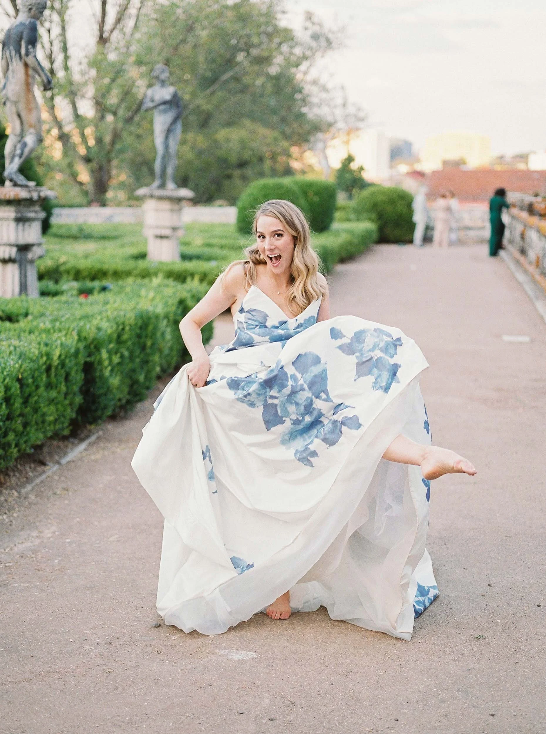 A young bride in a white dress with blue floral patterns posing playfully outdoors on a gravel path, surrounded by greenery and statues on Palácio dos Marqueses da Fronteira in Lisbon