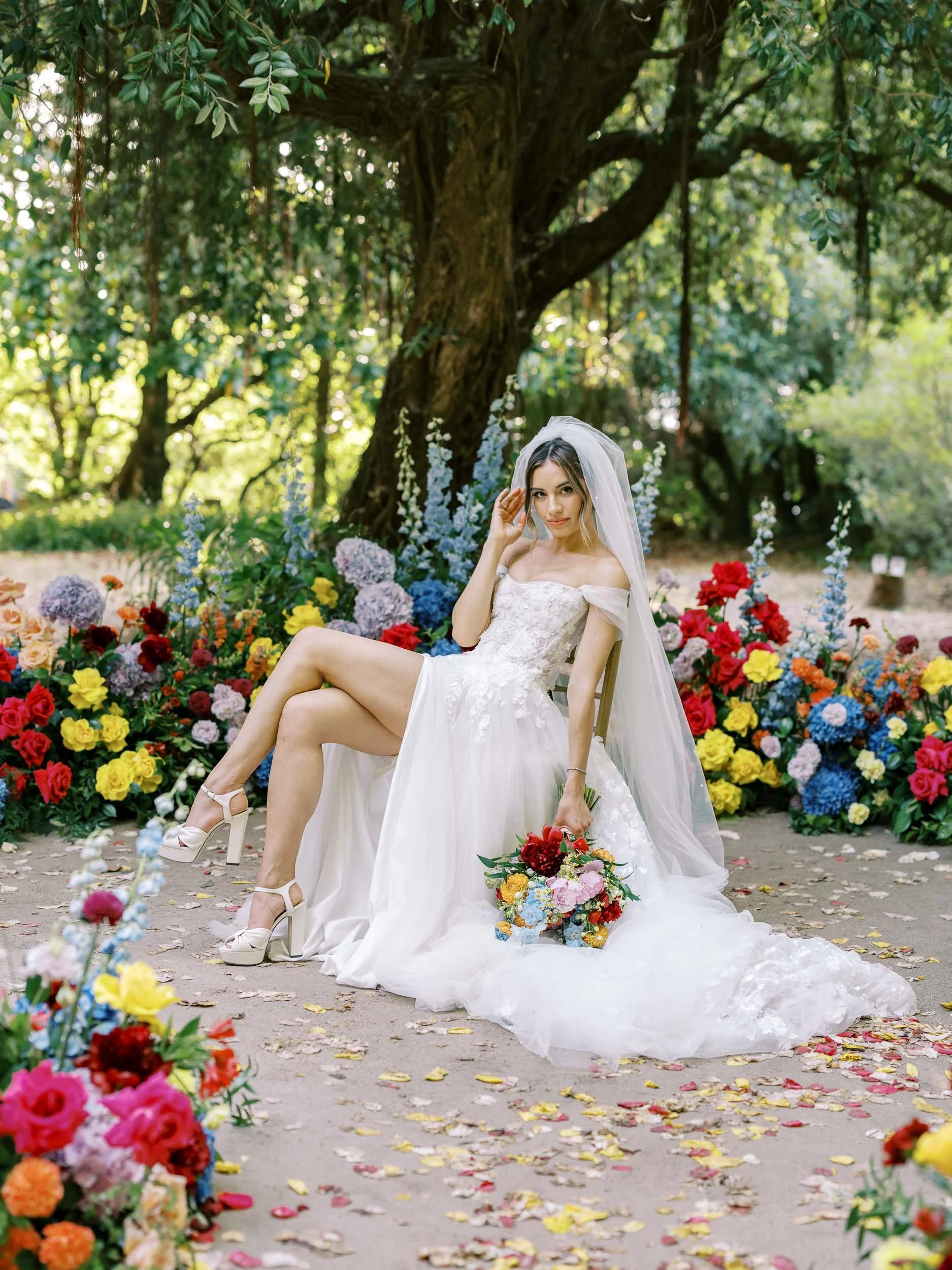 A bride in a wedding dress sitting outdoors under a large tree, surrounded by colorful flowers, holding a bouquet, and posing with one leg crossed over the other at Casa dos Penedos in Sintra