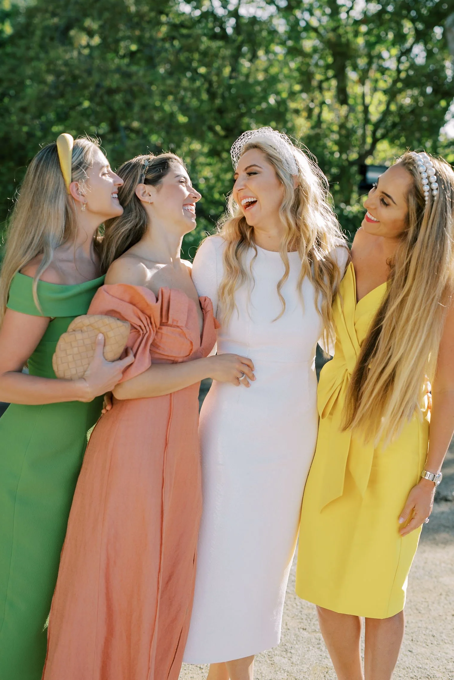 Bride and Bridesmaids smiling and laughing together outdoors, with green trees in the background. The bride is wearing a white dress and a veil, and the others are dressed in bright green, peach, and yellow dresses.