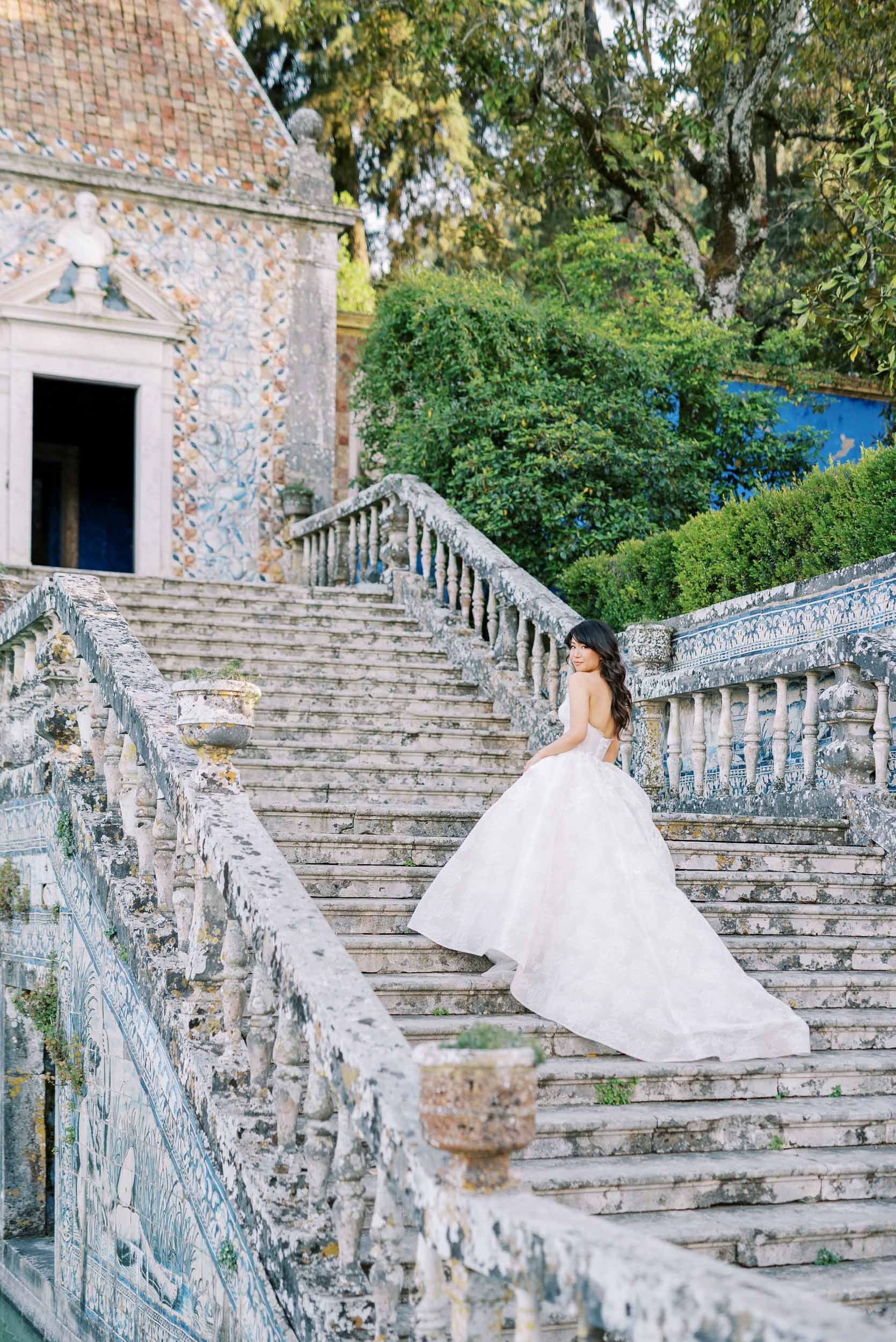 A woman in a strapless white wedding dress sitting on outdoor stone stairs with a lush garden and historic building in the background at Palácio dos Marqueses da Fronteira in Lisbon