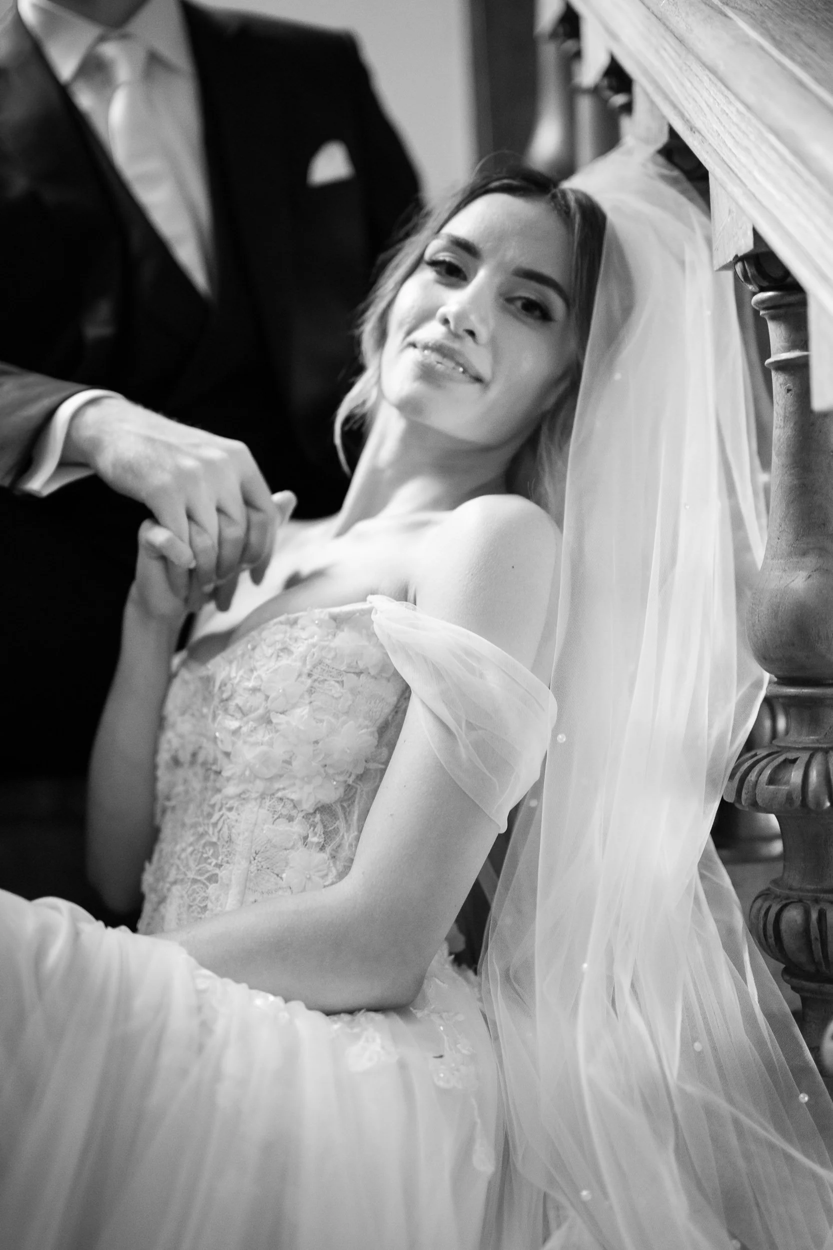 A black and white photo of a bride in a wedding gown with a veil, leaning against a bannister, smiling while holding hands with a groom dressed in a tuxedo.