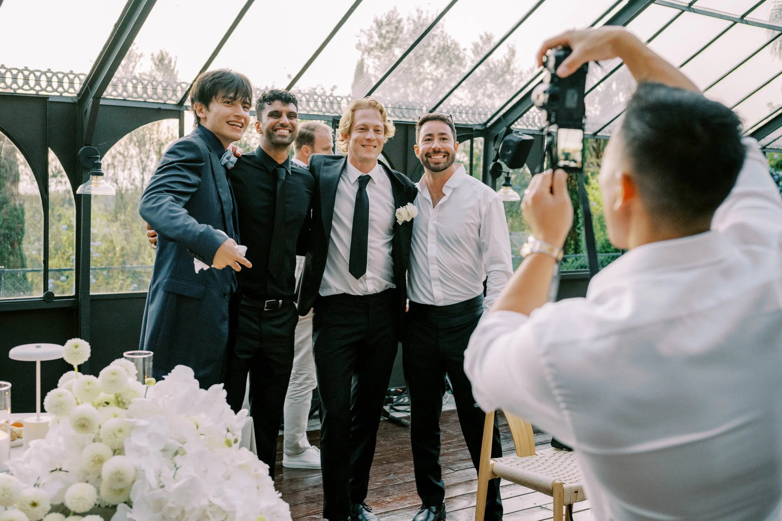 Four wedding guests in formal attire posing for a photo at a wedding or celebration in a glass-roofed venue. One man has a white flower boutonniere. A person in the foreground is taking the picture with a camera at Quinta da Bella Vista