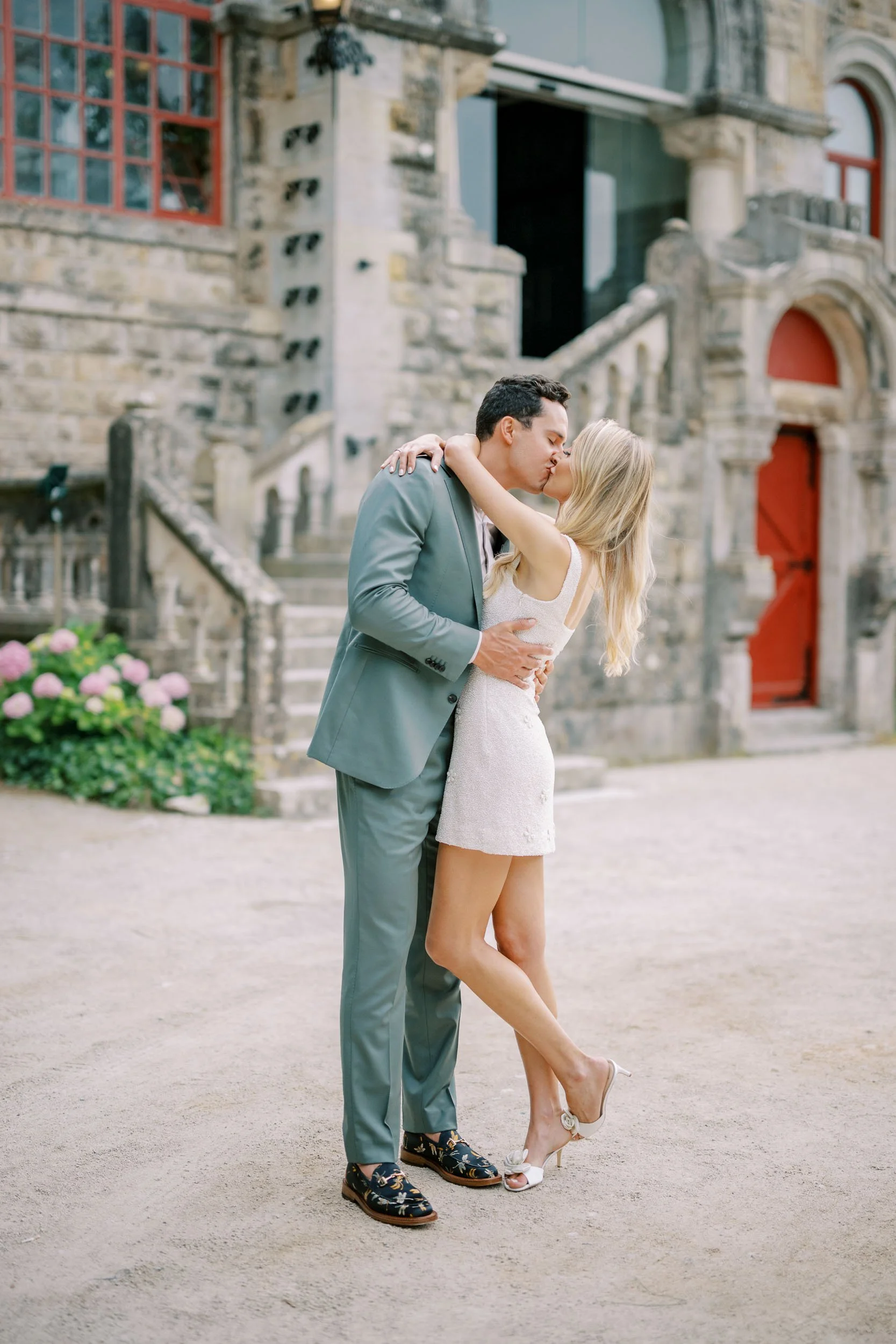 A man and woman kissing outdoors in front of a historic stone building with stairs and red doors.