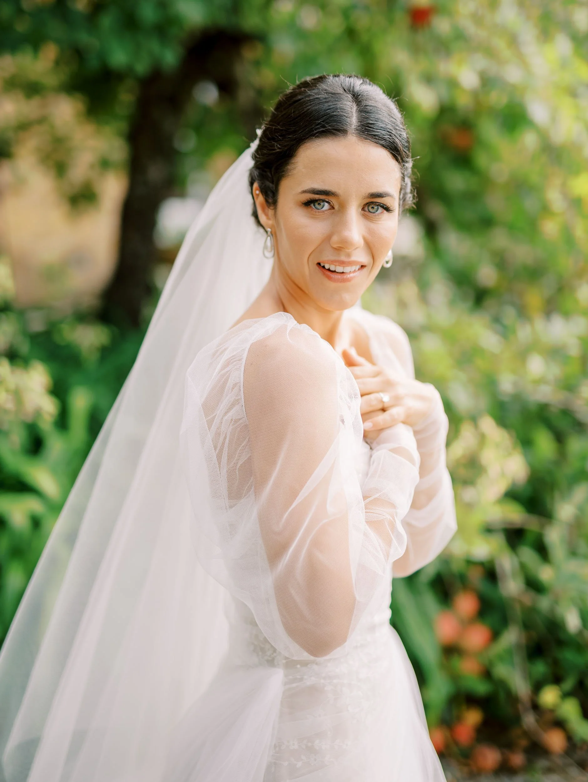 Bride with dark hair in a wedding dress and veil, outdoors in a garden with greenery, looking at the camera with a gentle smile.