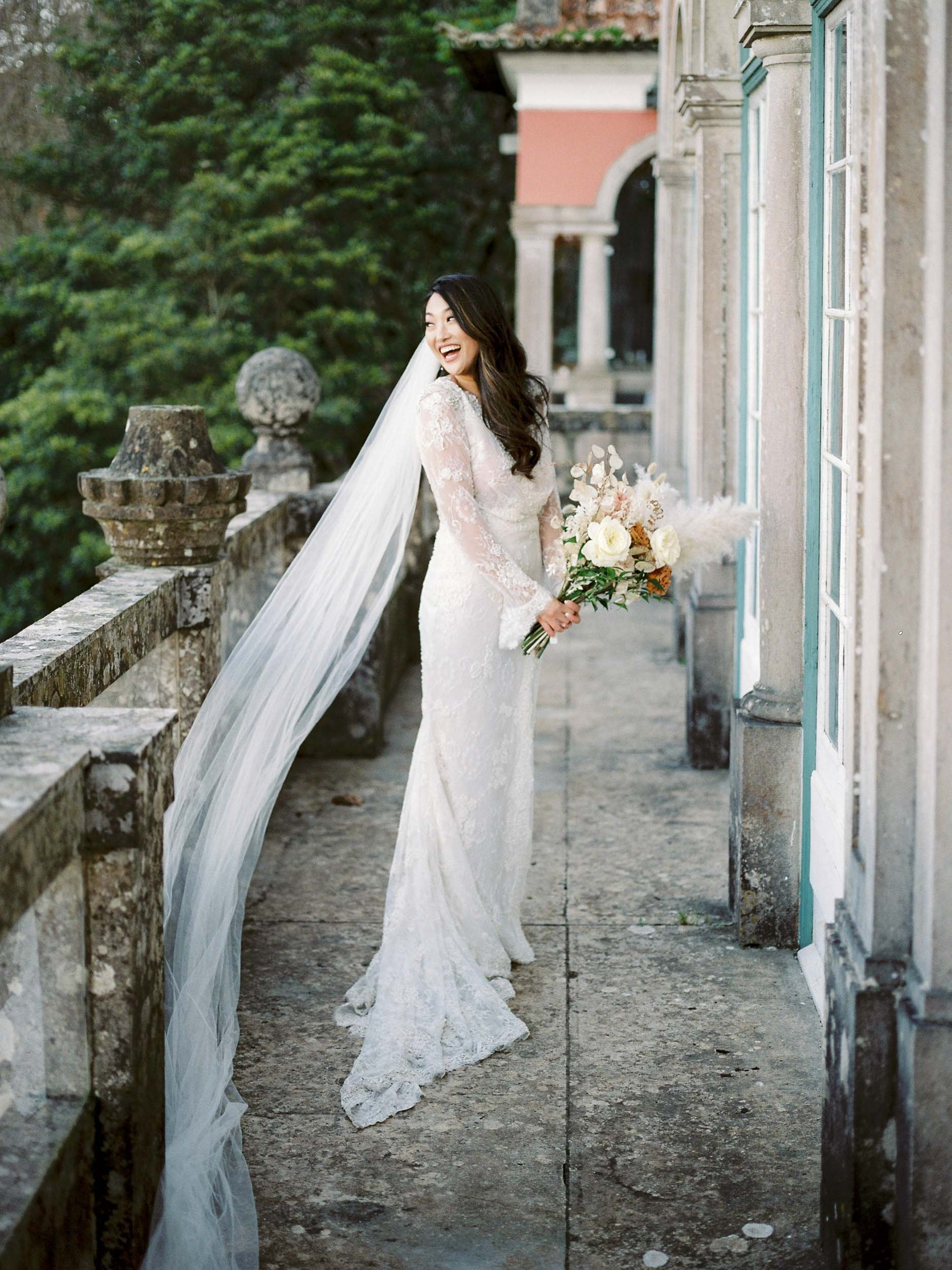 Bride in a long white lace wedding dress with a flowing veil holding a bouquet of white and pastel flowers, smiling on a stone balcony surrounded by lush greenery and historic architecture at Casa dos Penedos in Sintra, Portugal