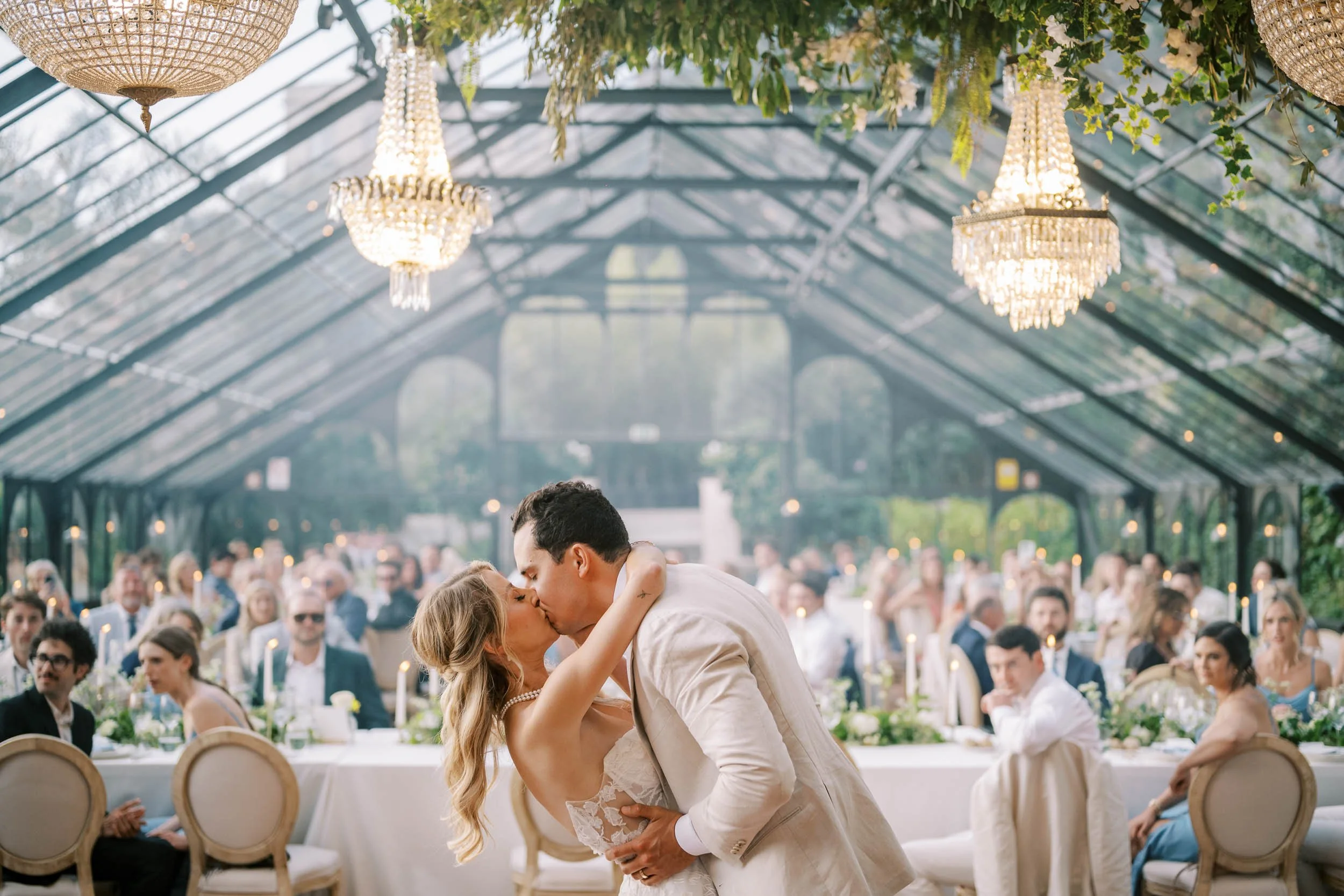 A newlywed couple sharing a kiss at their wedding reception in the glass house of Quinta da Bella Vista with chandeliers and seated guests in the background.