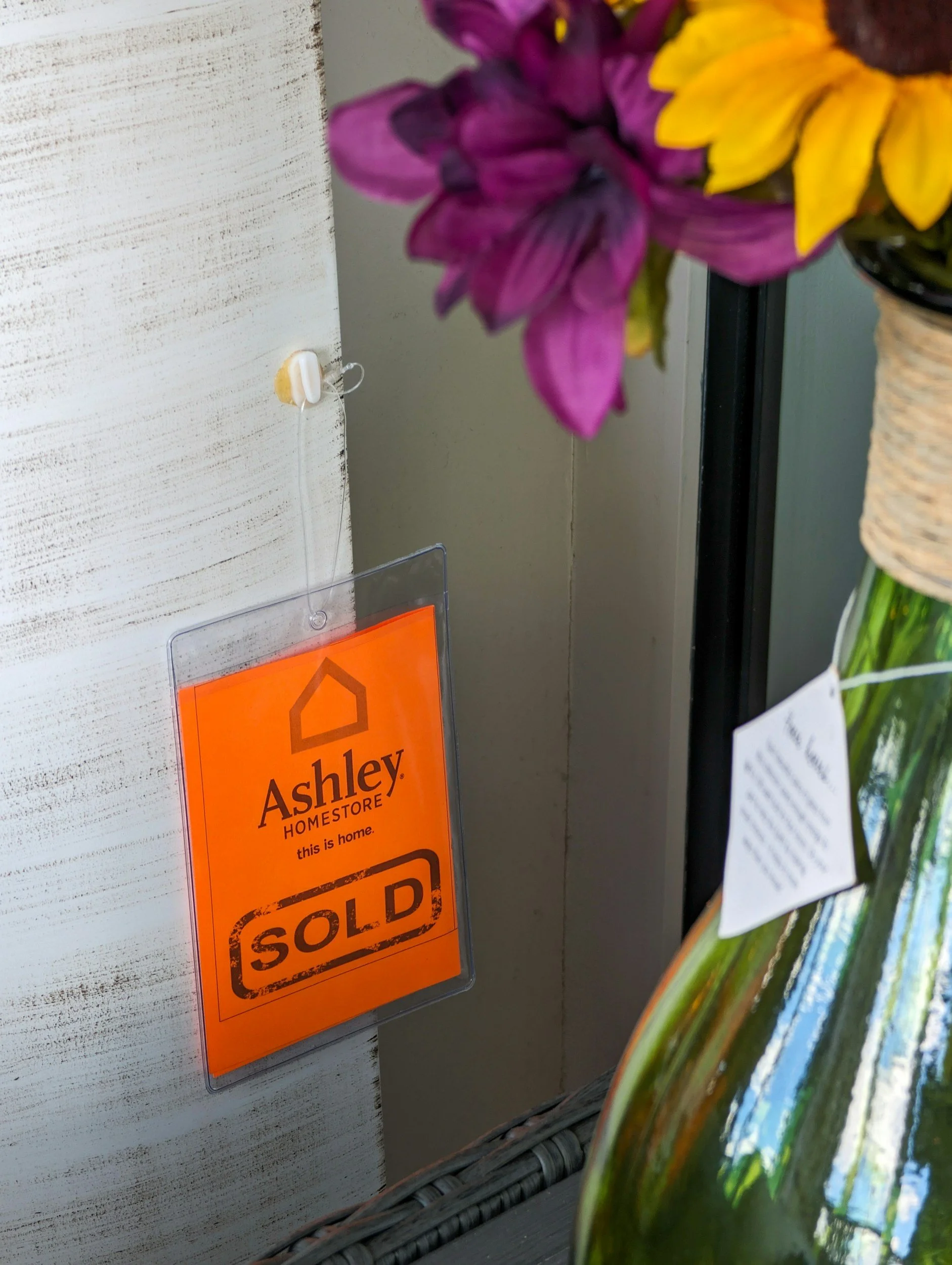 Close-up of a wall with an orange 'Sold' sign for Ashley HomeStore, partially obscured by a colorful ceramic vase with flowers, including purple petals and a yellow center.