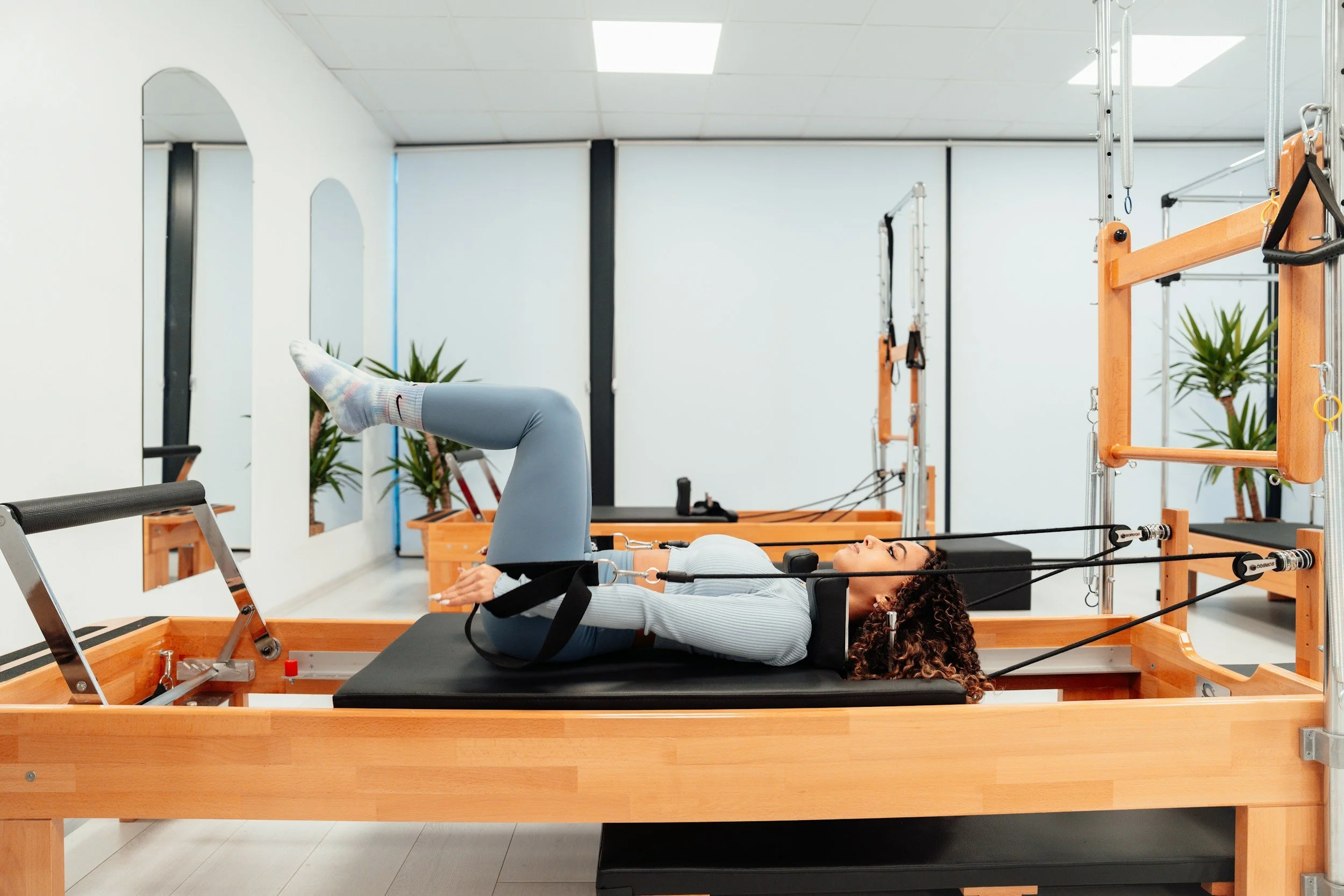 A woman with curly hair lying on a Tower reformer machine, exercising her legs while holding straps in a bright fitness studio with motivational posters and plants.