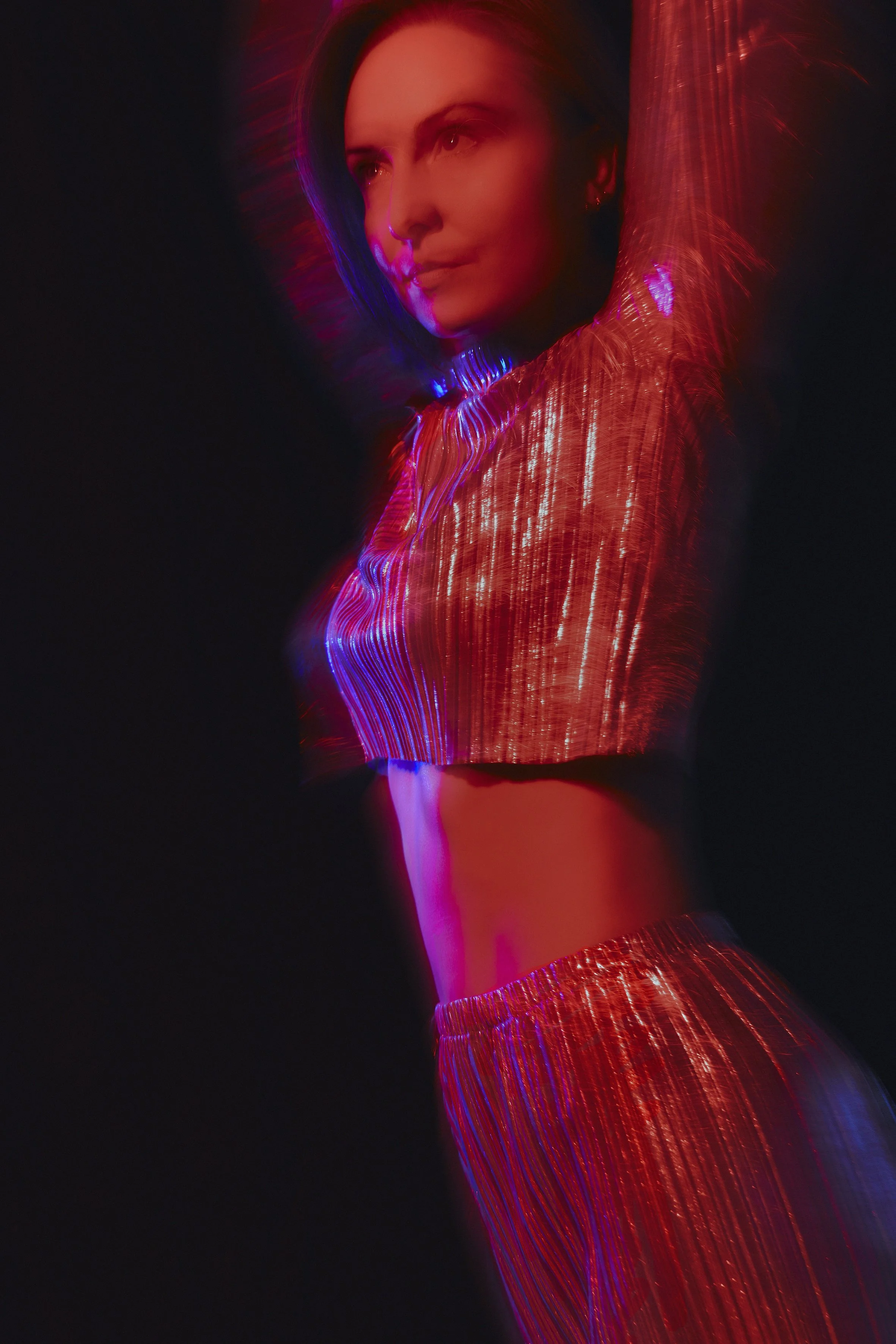 A woman in a shiny, metallic red two-piece outfit with a crop top and pants, posing with her arm raised against a dark background with colorful light effects.