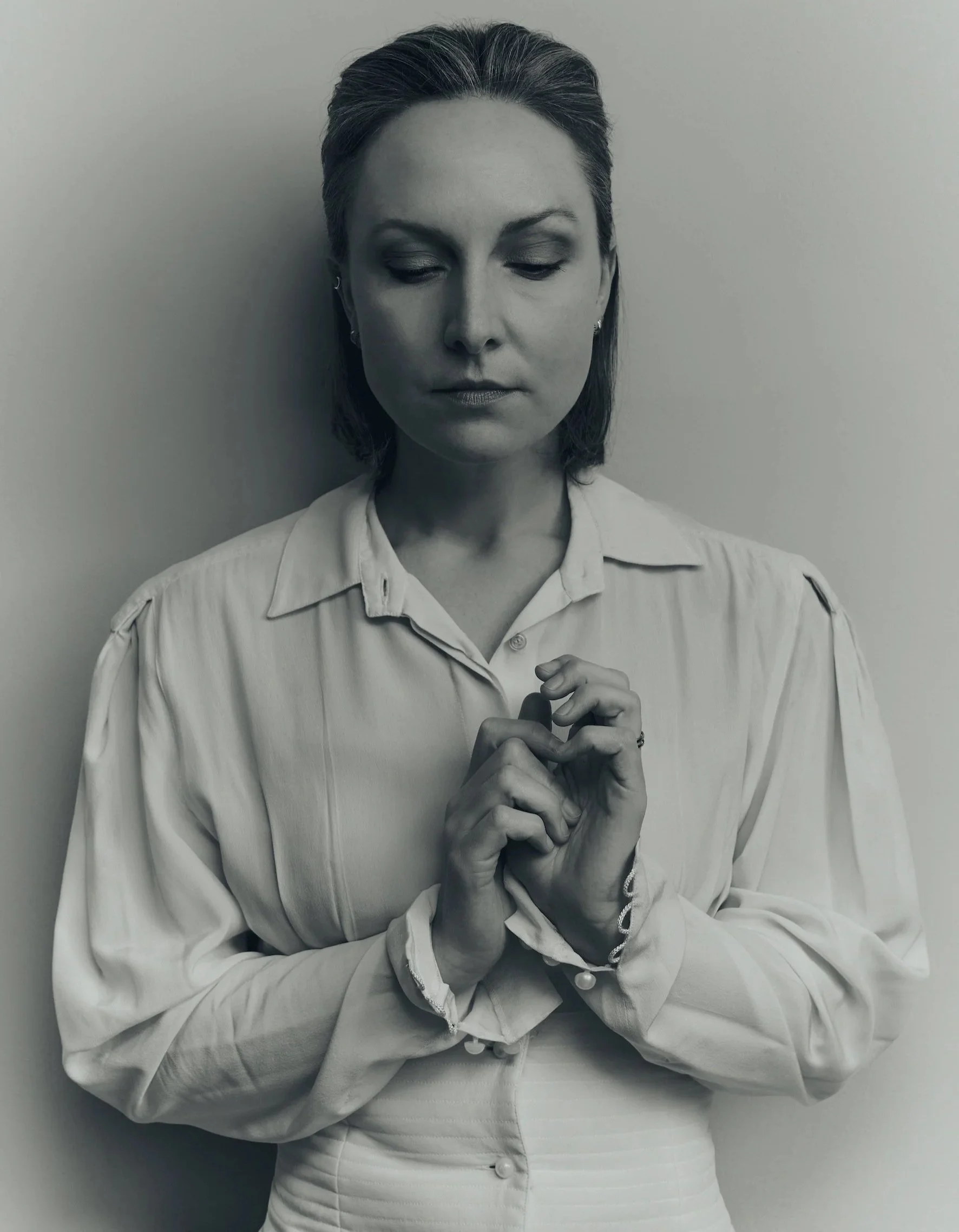 Black and white photo of a woman with short hair, eyes closed, holding her hands together near her chest, standing against a plain background.