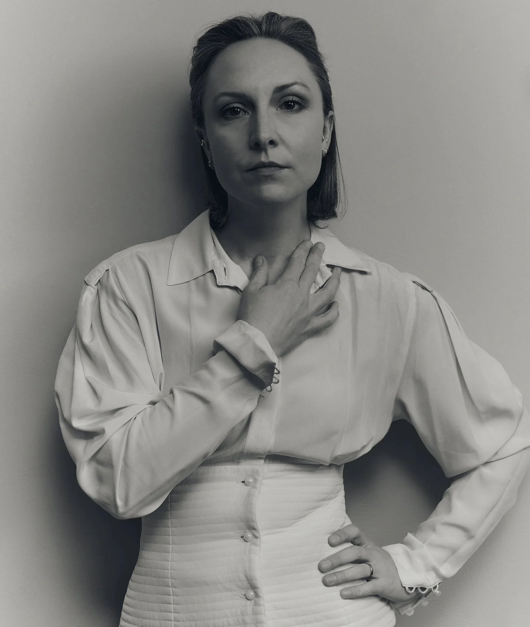 Black and white photograph of a woman wearing a vintage long-sleeve blouse and a high-waisted skirt, with her hand on her chest and the other on her hip, standing against a plain background.