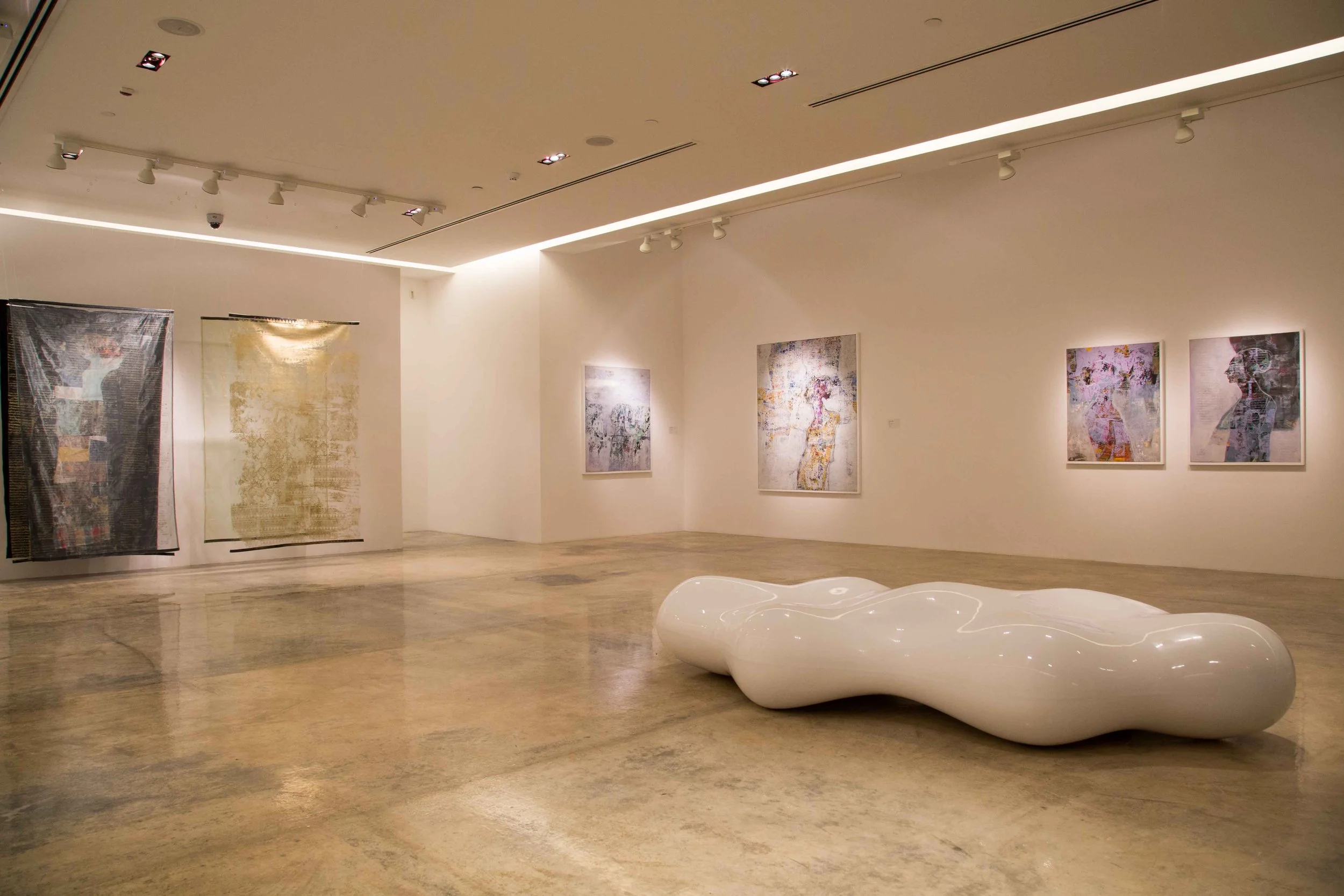 An art gallery with white walls displaying abstract paintings. A large, irregularly shaped white sculpture is in the foreground on a polished concrete floor.