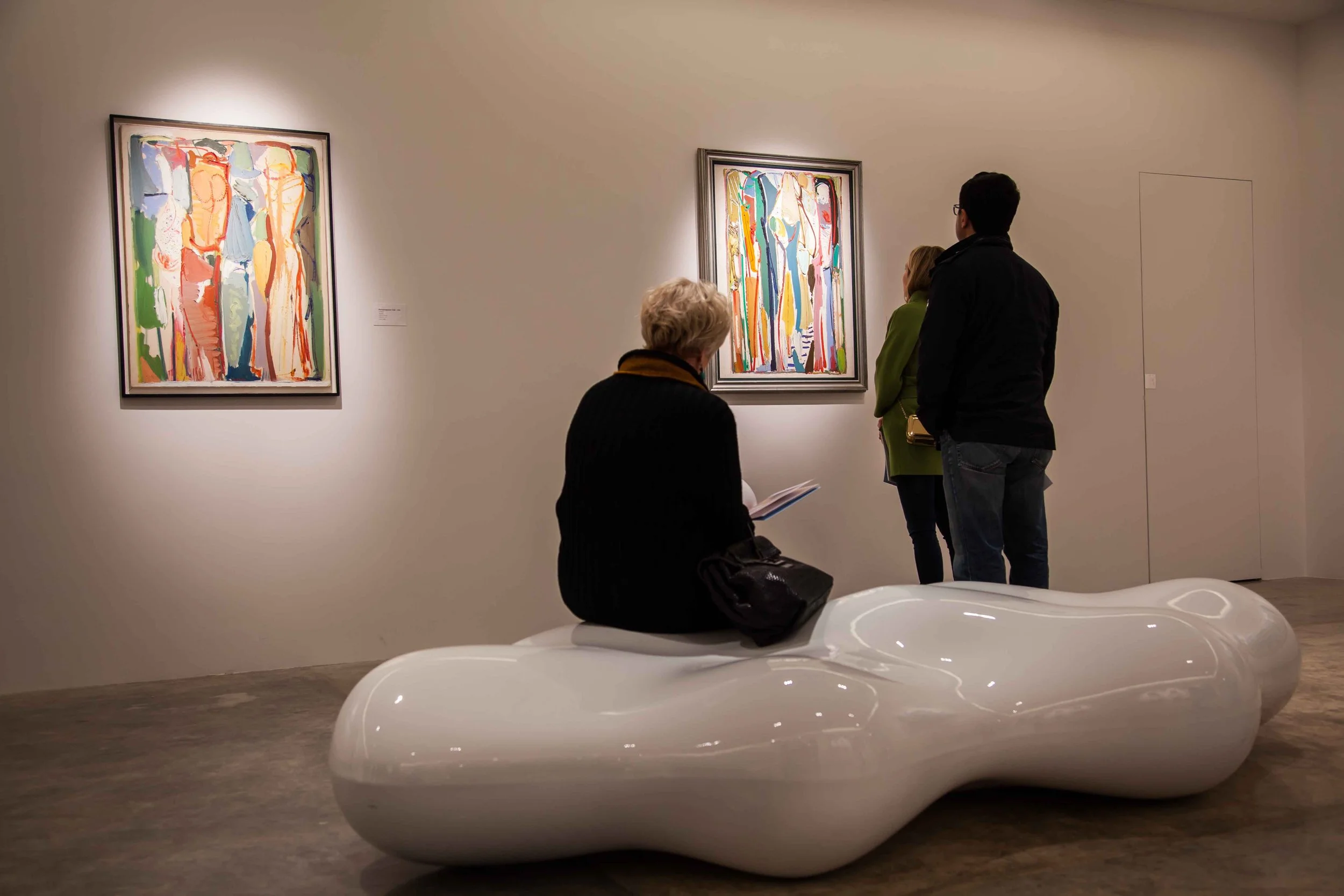 People viewing colorful abstract paintings in an art gallery, with one person sitting on a modern, white, curved bench.