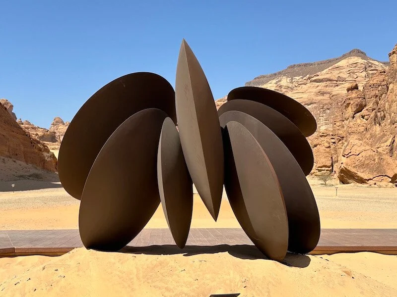 A modern abstract sculpture made of large dark metal oval and circular shapes, set against desert landscape with rocky hills and clear blue sky.