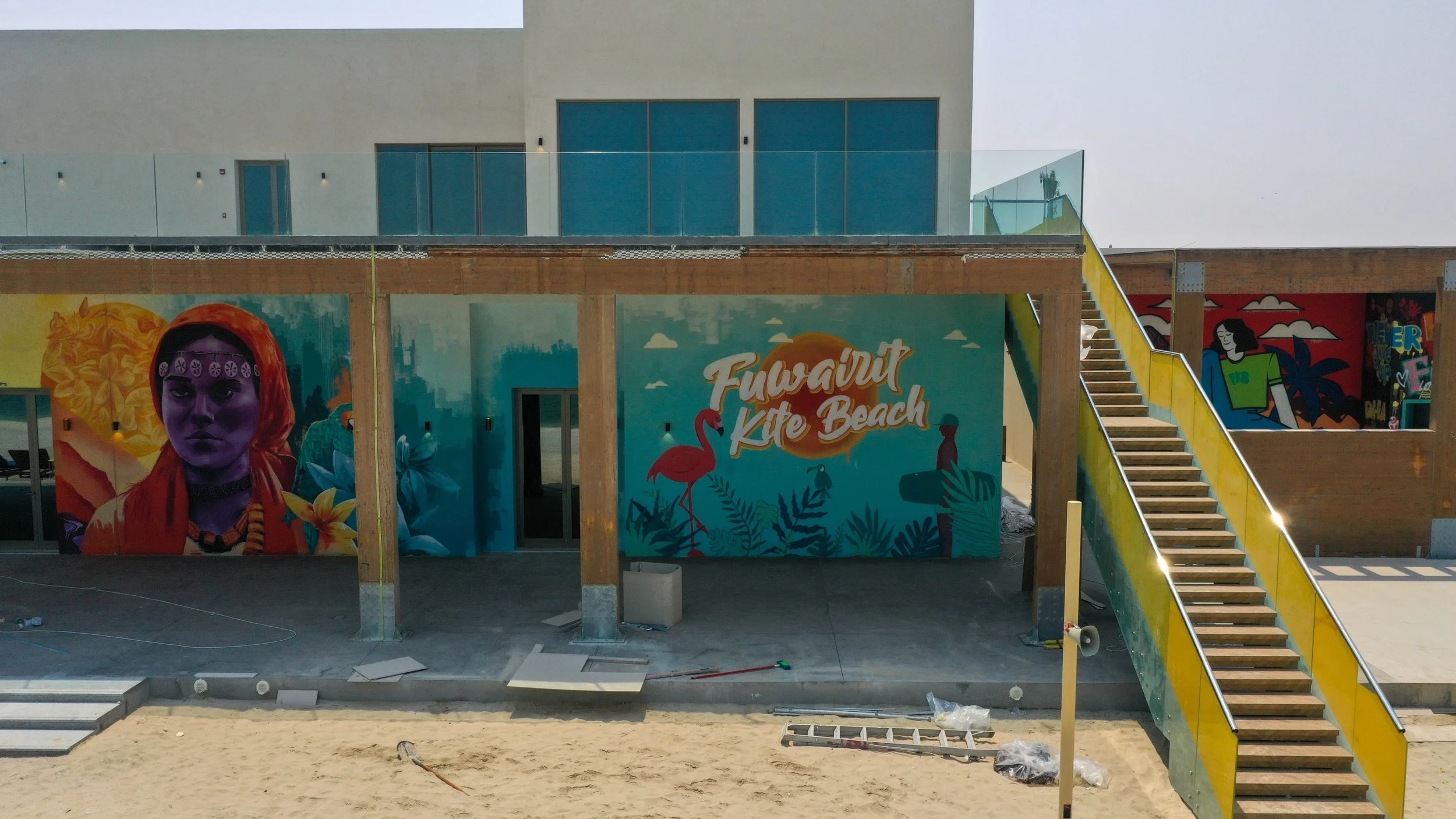 A colorful mural on a building displaying the words "Fuweint Kite Beach," with a pink flamingo, lush tropical plants, and a person silhouette, with additional murals visible on the right side of the image.