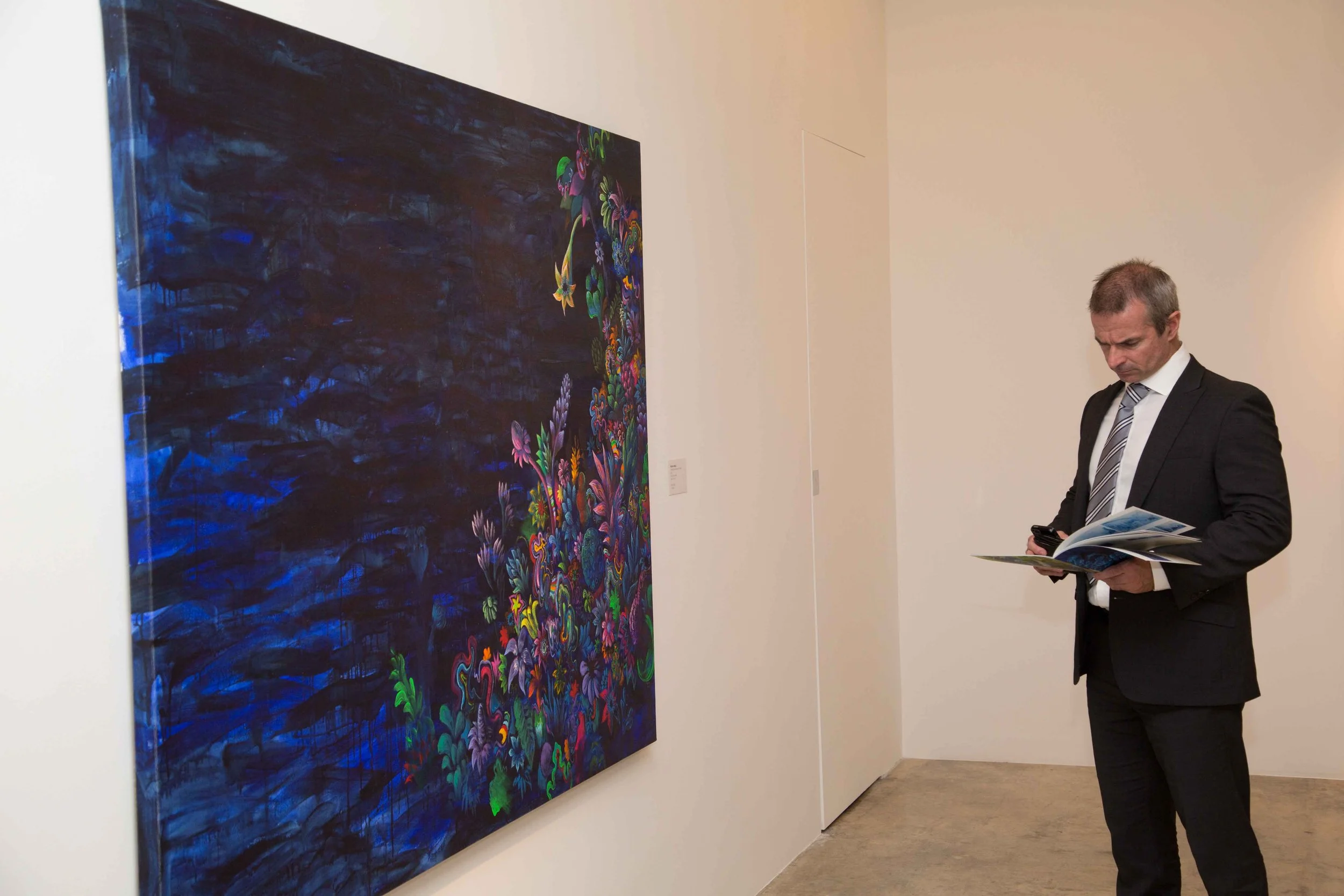 A man in a black suit and striped tie is looking at a gallery wall while holding a brochure and a smartphone.