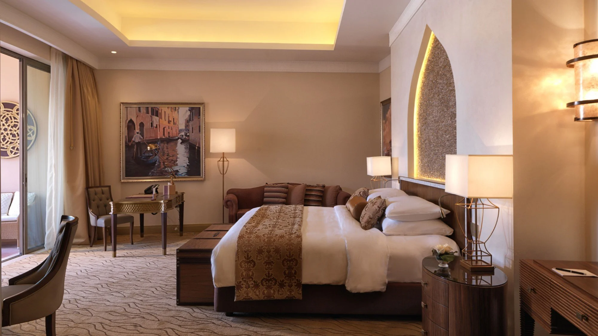 Interior of a luxurious hotel room featuring a king-sized bed with white linens, decorative pillows, and a gold and brown runner. To the right of the bed, there are two bedside lamps on wooden nightstands, and above the bed, a textured wall niche wit