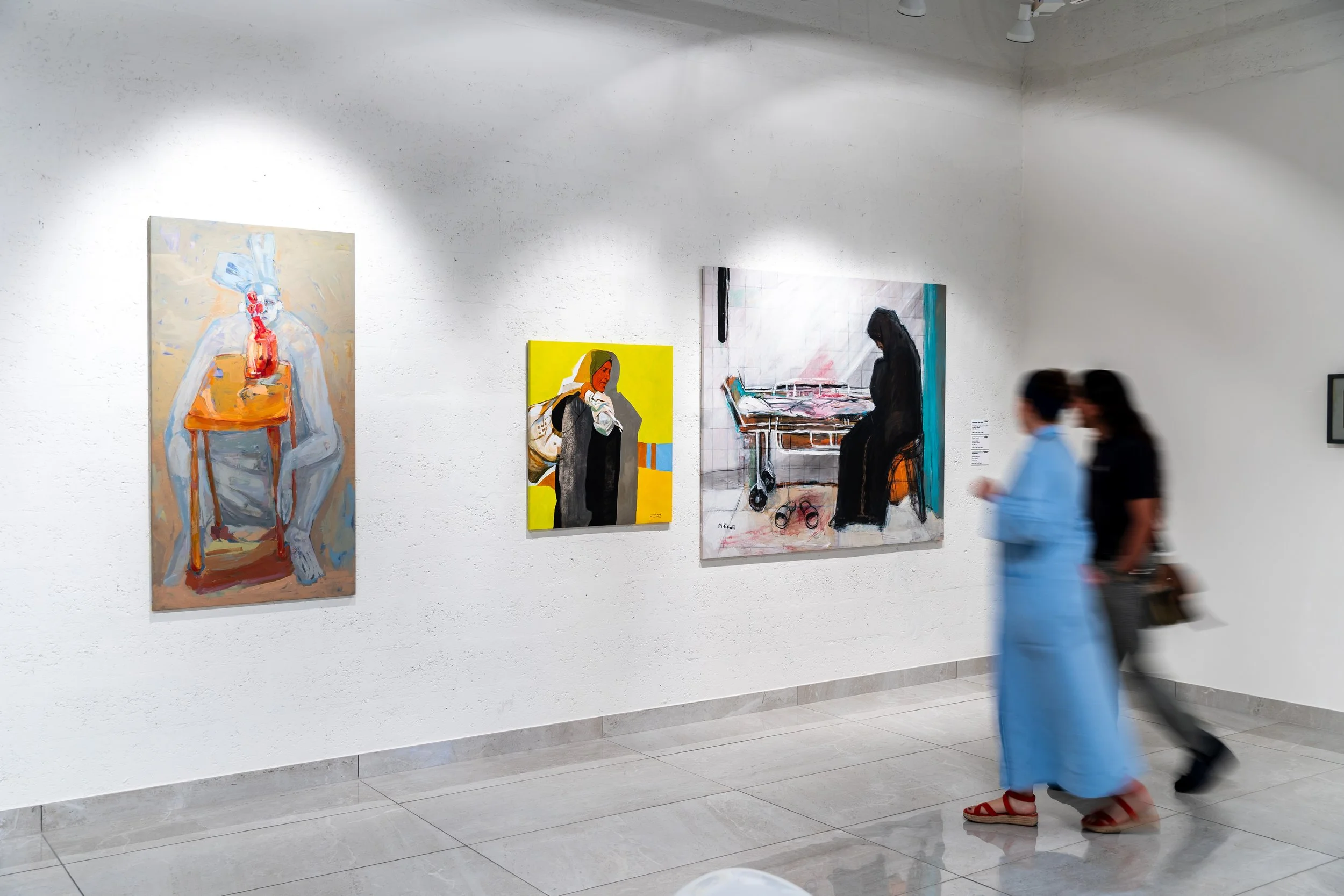 People walking past paintings displayed on a white gallery wall.