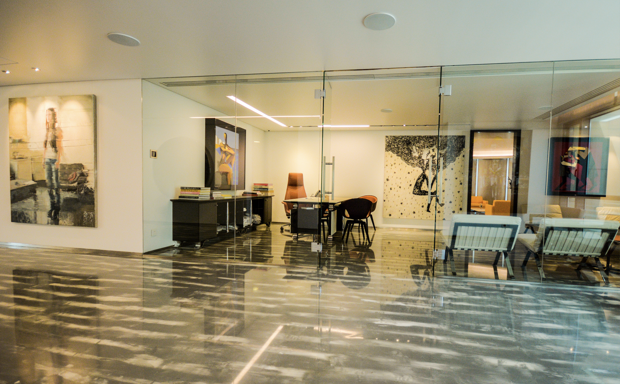 Modern office space with a glass wall, wooden flooring, and artwork on the walls, containing a desk, chairs, and shelves.