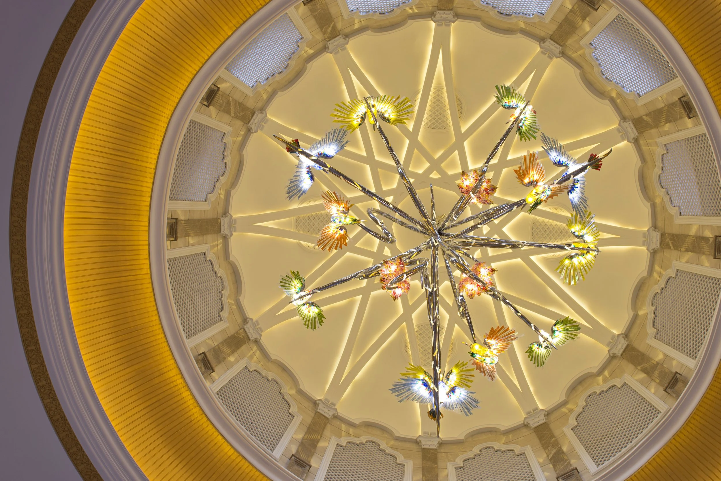 Colorful modern chandelier with illuminated, multicolored glass or plastic leaf-like shapes hanging from a shiny metal structure, seen from below inside a building with a domed ceiling and a yellow decorative circular border.