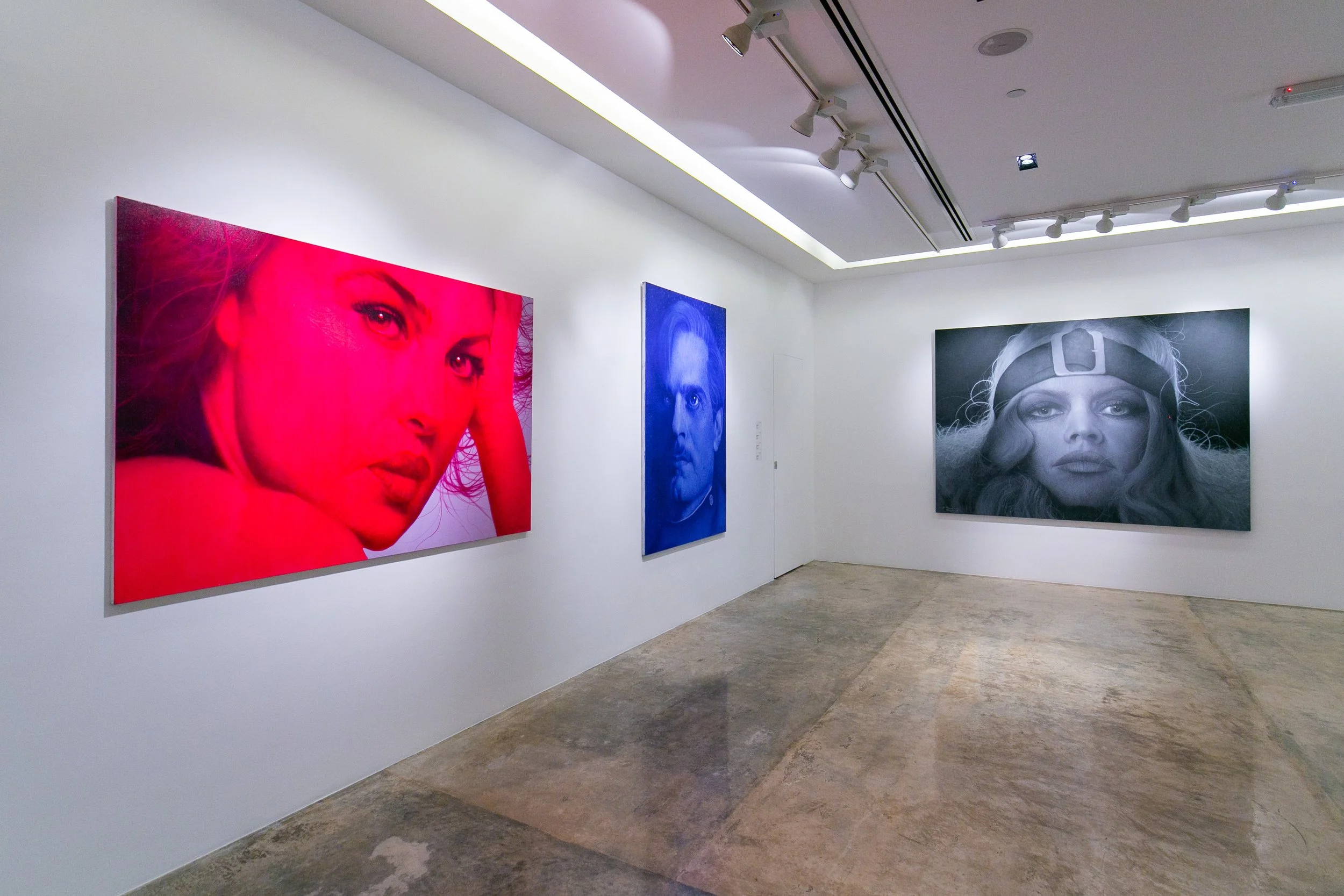 Gallery with three portrait paintings on white wall, featuring a woman with red lighting, a man with blue lighting, and a woman with black and white tones, in a modern art gallery.