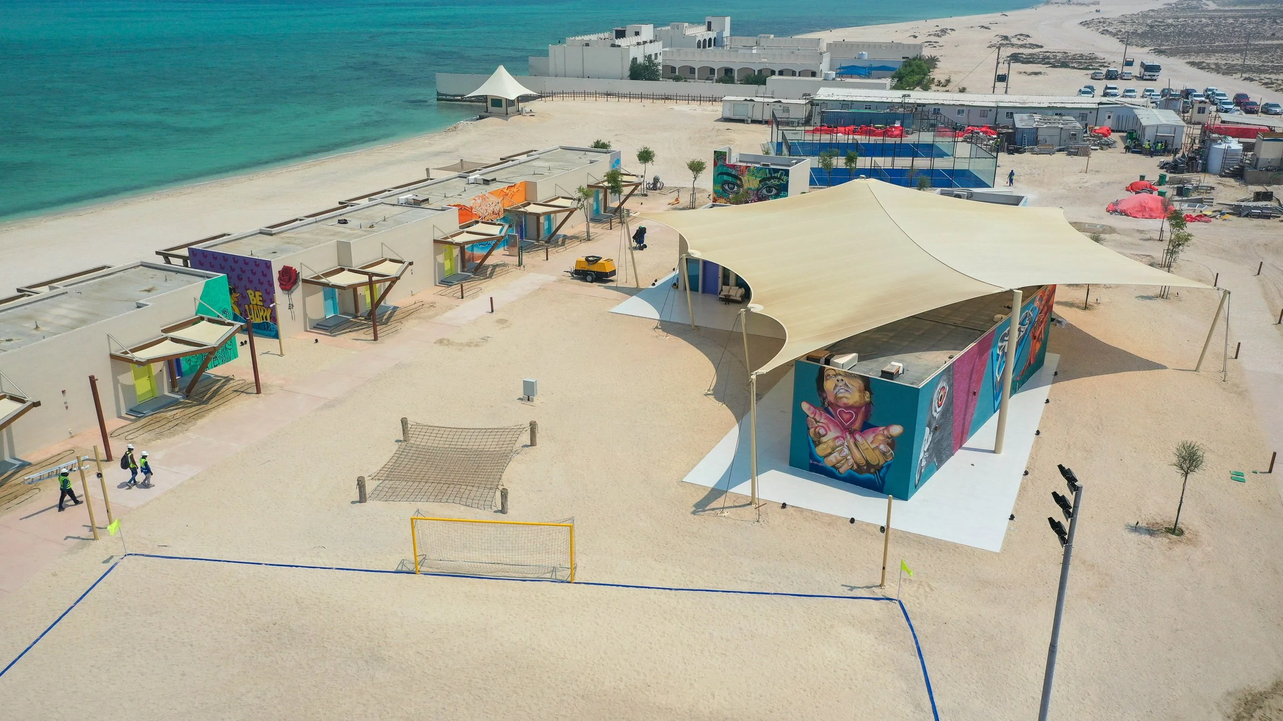 Aerial view of a beach with volleyball courts, a stage with a canopy, small huts, and art murals on buildings near the water.