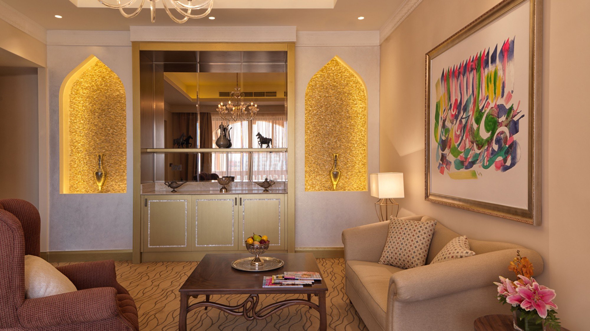 A luxurious living room with beige walls, a painting on the right wall, a beige sofa with patterned pillows, a small wooden table with magazines and a fruit bowl, a pink flower vase on a side table, and decorative alcoves with gold backlighting displ