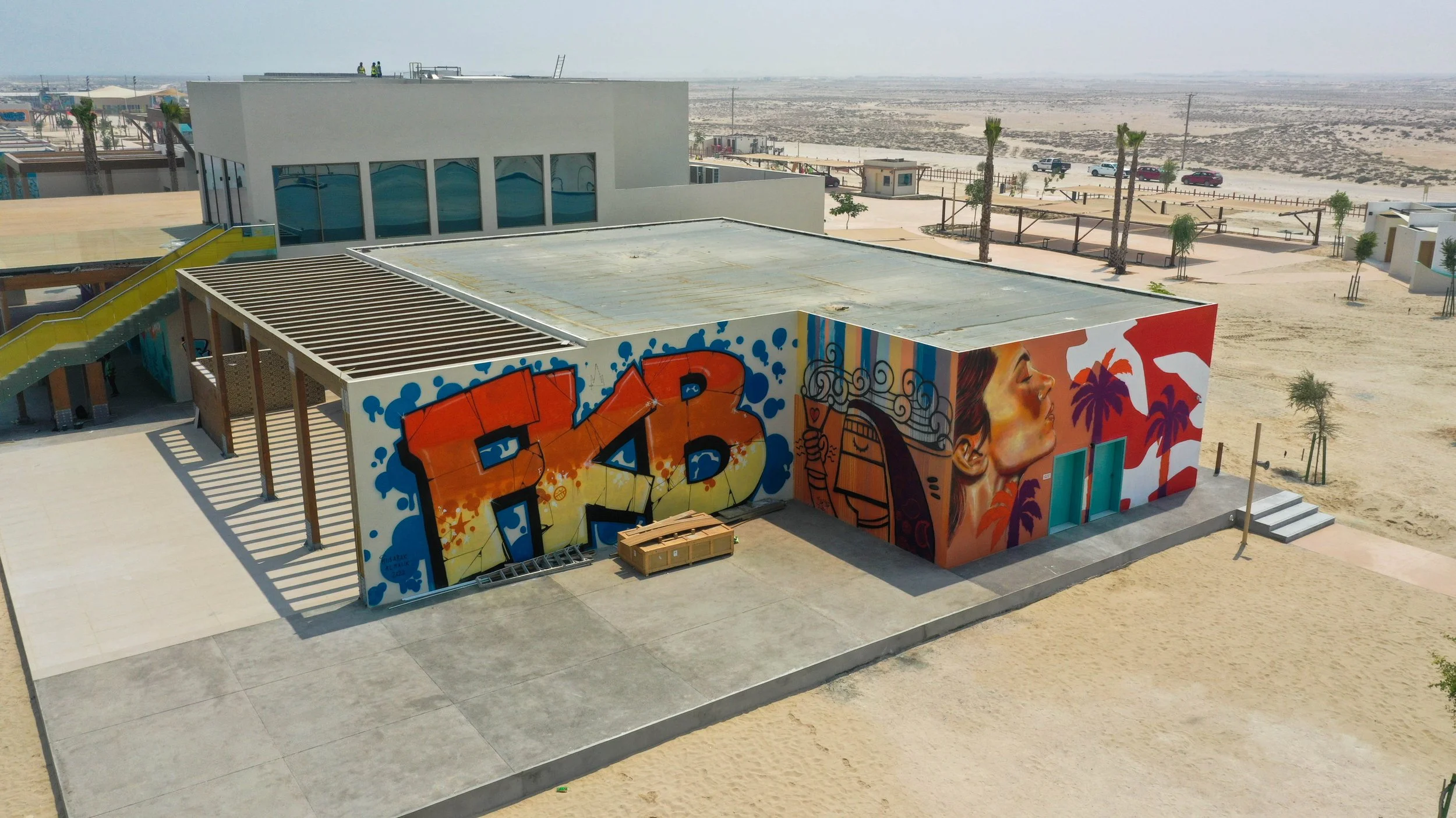 A building with colorful graffiti art on its walls, including large 3D letters and a woman's face with palm trees and sunset background. Desert landscape in the background.