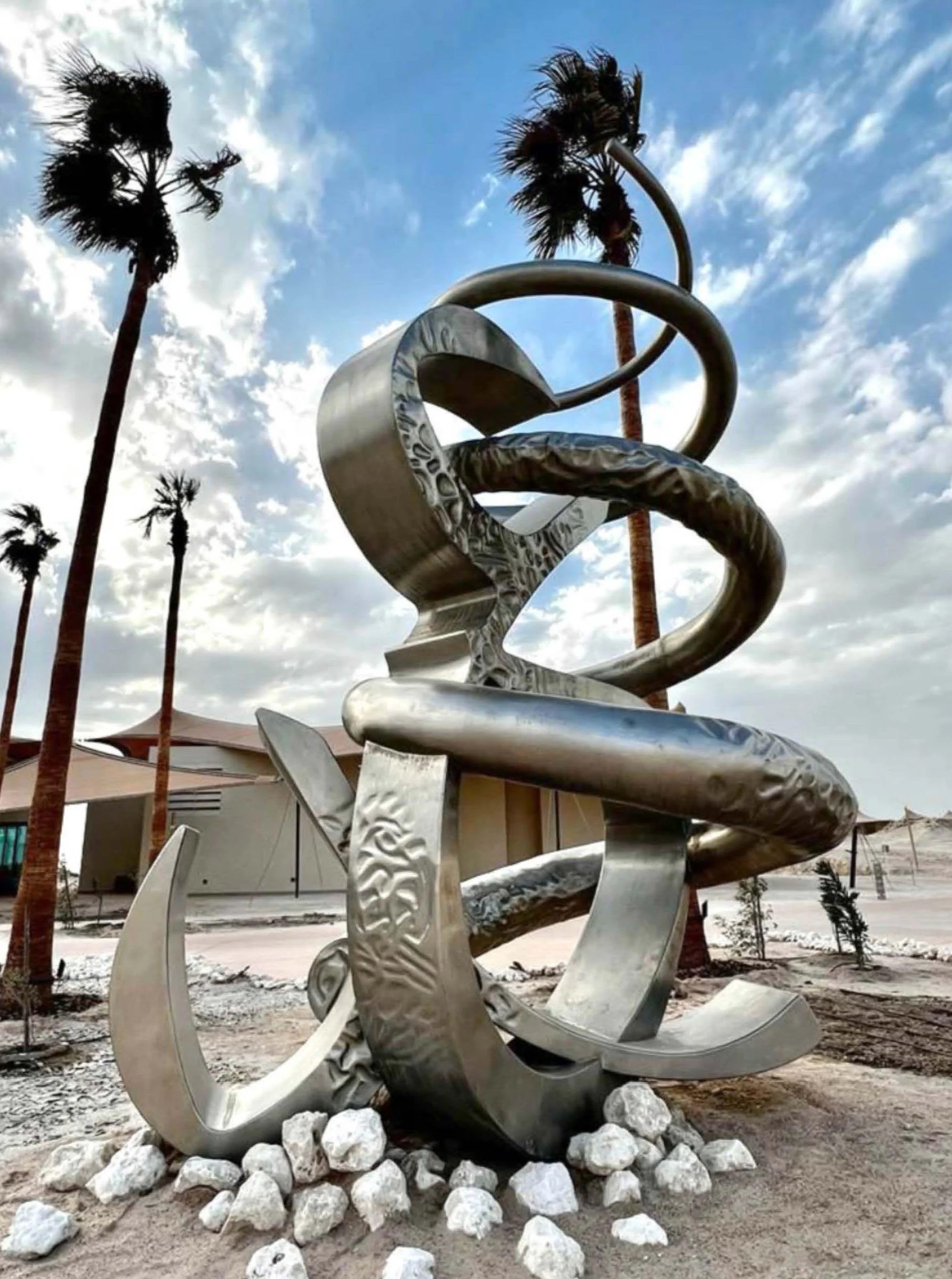 Large modern metal sculpture with abstract, swirling shapes, resting on white rocks, surrounded by tall palm trees, in an outdoor setting with a partly cloudy sky.