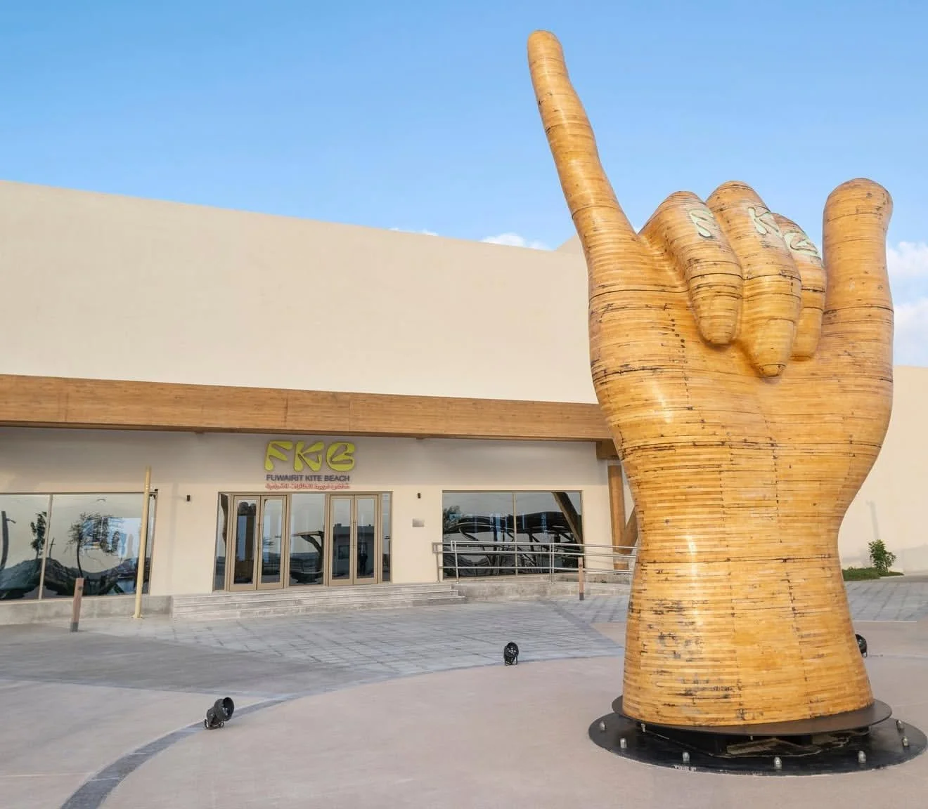 A large wooden sculpture of a hand making the 'I love you' sign in sign language outside a building with a sign that reads 'Funtner Kite Beach'.