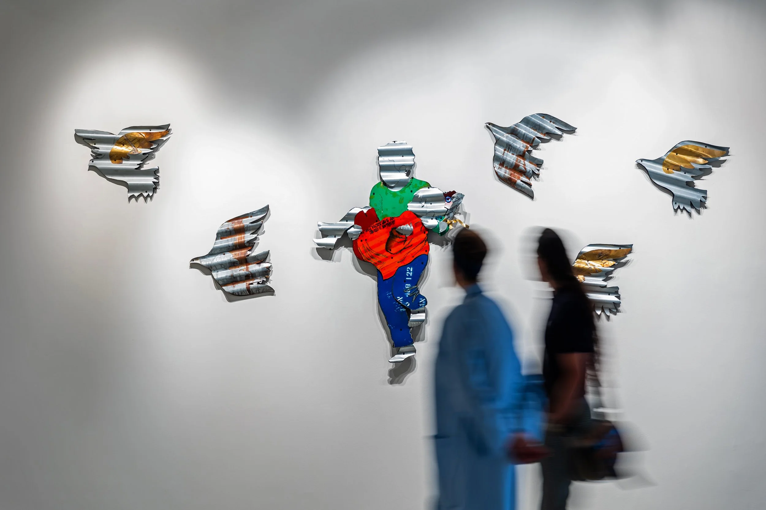 Contemporary art installation of a colorful runner figure made from metal, surrounded by metal bird cutouts, displayed on a white gallery wall, with two blurred people walking past in the foreground.