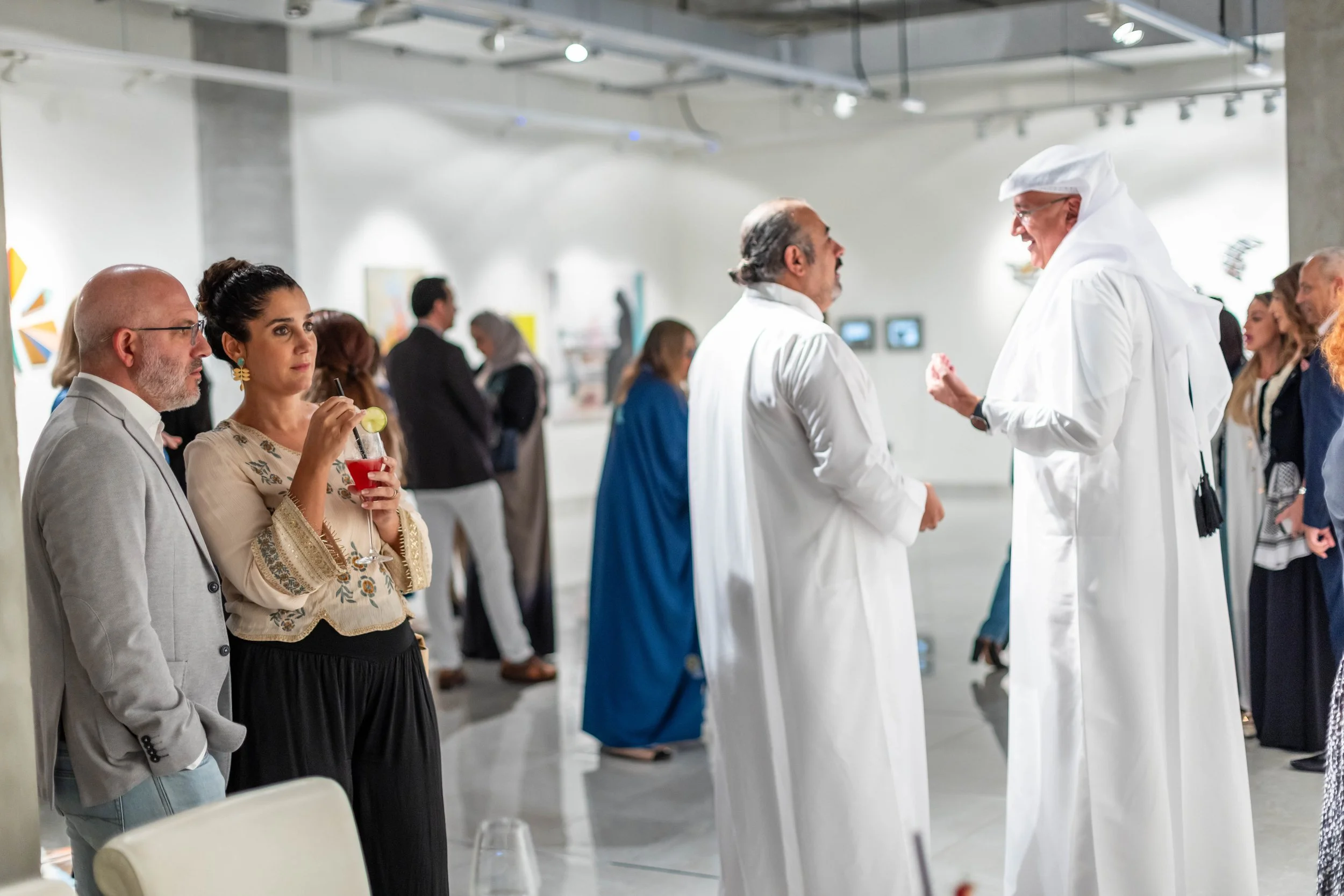 People attending an art gallery or museum, some engaging in conversations while others enjoy drinks; the setting is modern with white walls and artworks on display.