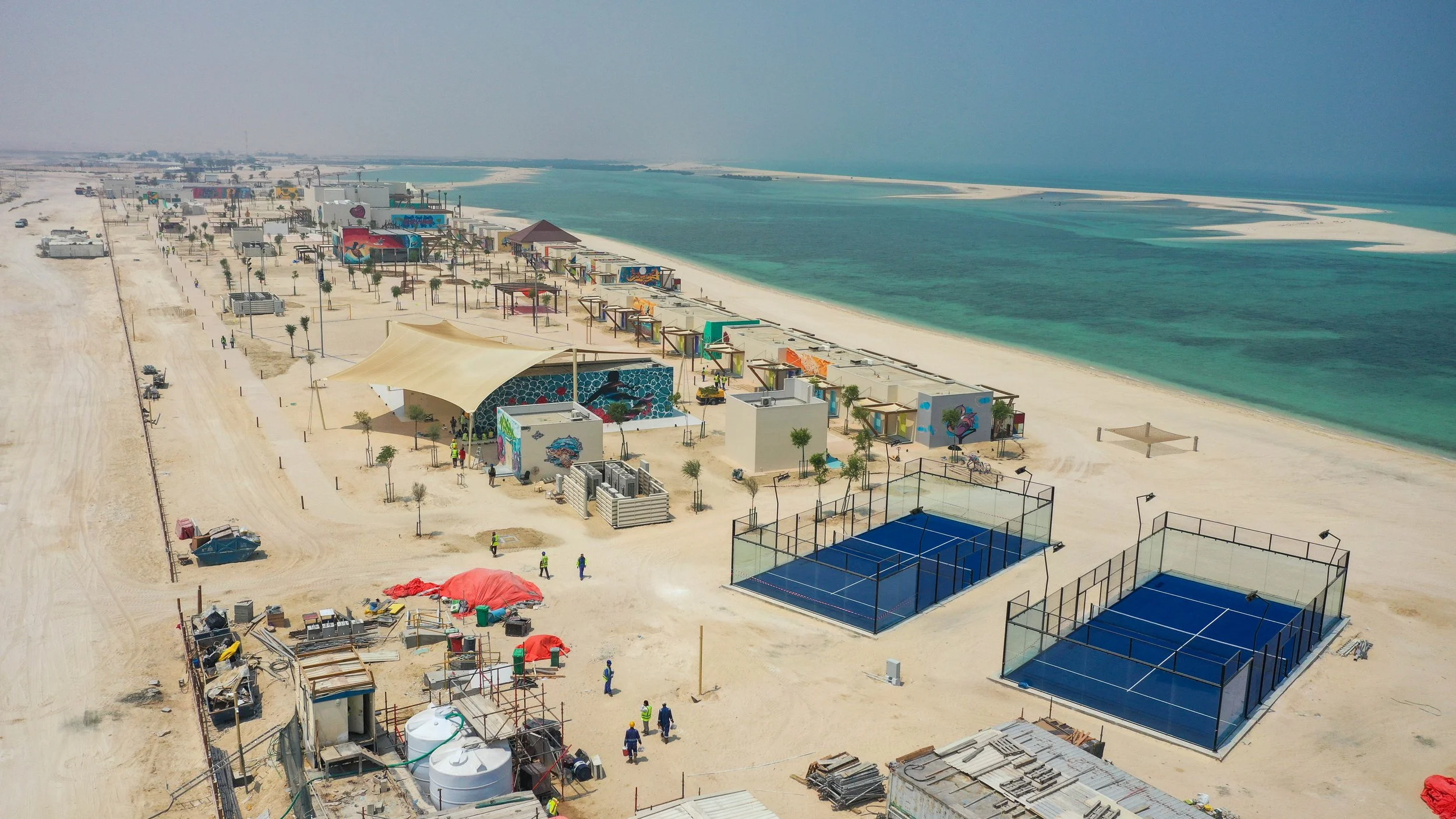 Aerial view of a beachside tourist area with colorful buildings, tennis courts, and construction activity near the shoreline, next to turquoise waters.