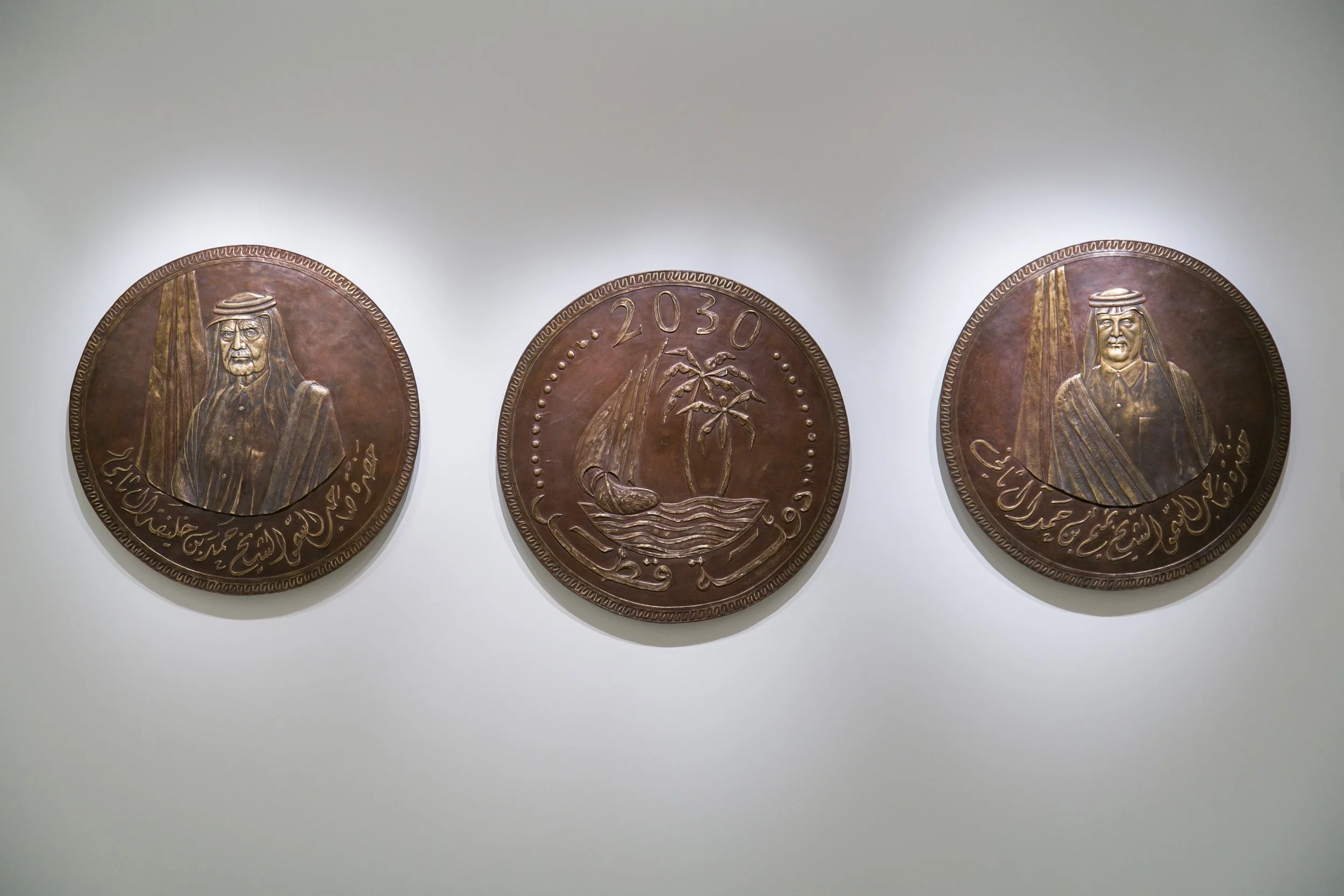 Three large, circular bronze plaques with embossed images and inscriptions mounted on a white wall. The plaques feature portraits of two men in traditional Middle Eastern attire, and a central emblem of a palm tree and a sailboat, with the years 2030