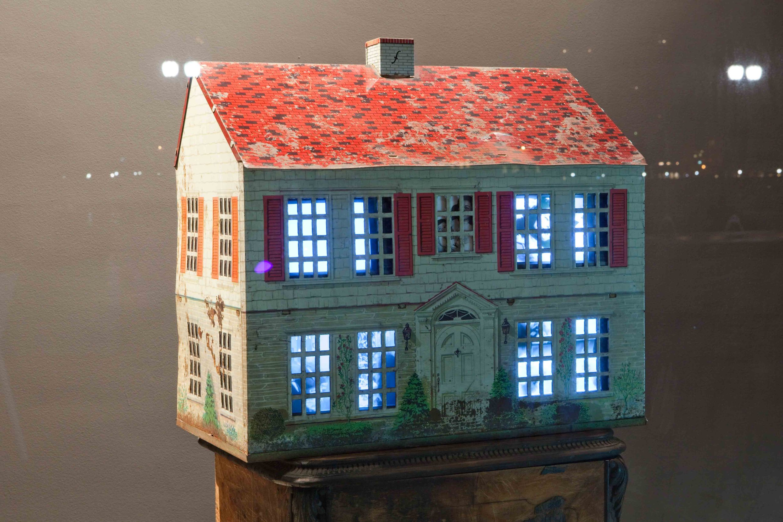 A small, illuminated, model house with a red shingled roof, white brick exterior, and multiple windows with purple and blue shutters. The house is placed on a wooden surface with a plain background.