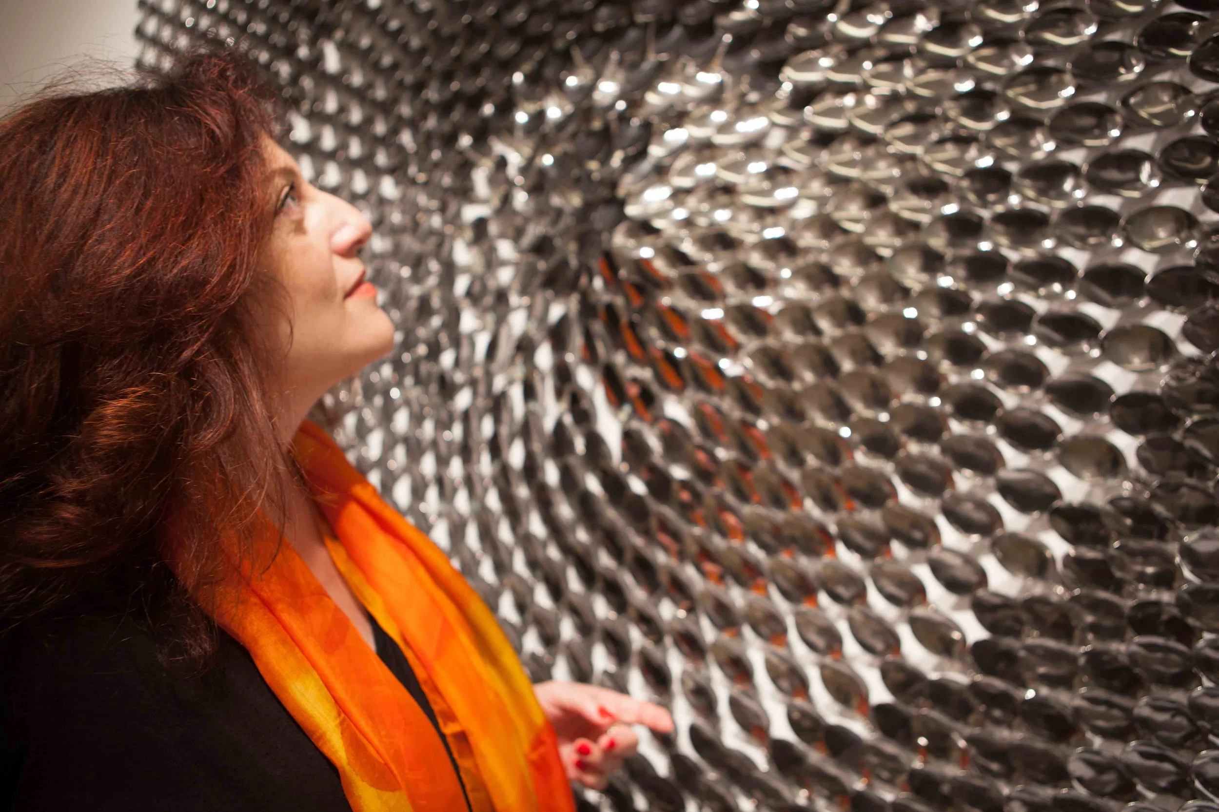 A woman with long red hair looking at a large wall of shiny metal bubbles or discs, wearing an orange scarf.