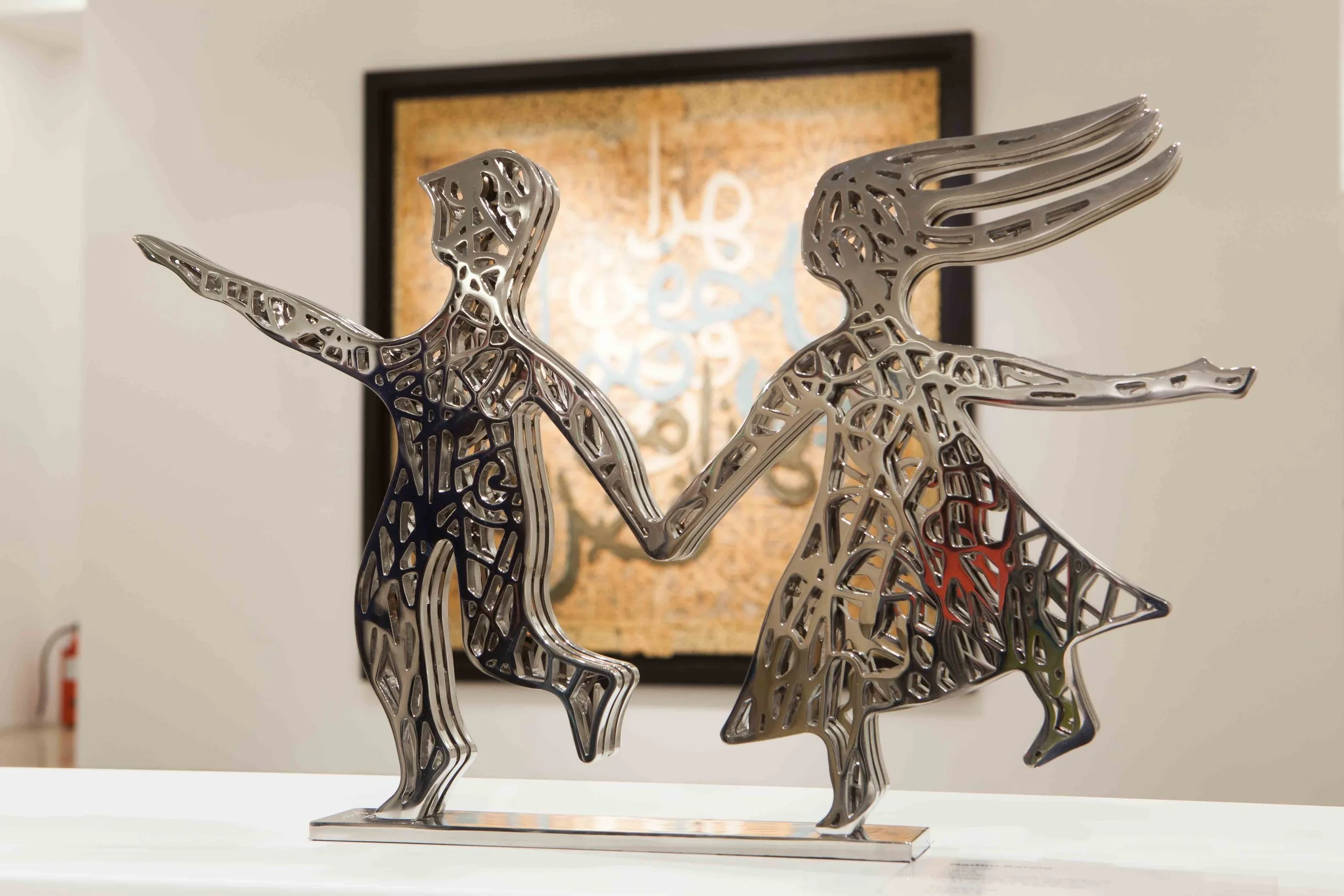 Metallic art sculpture of two stylized animals holding hands, one resembling a unicorn and the other a horse, with intricate cutout patterns, placed on a flat surface in front of a framed artwork.