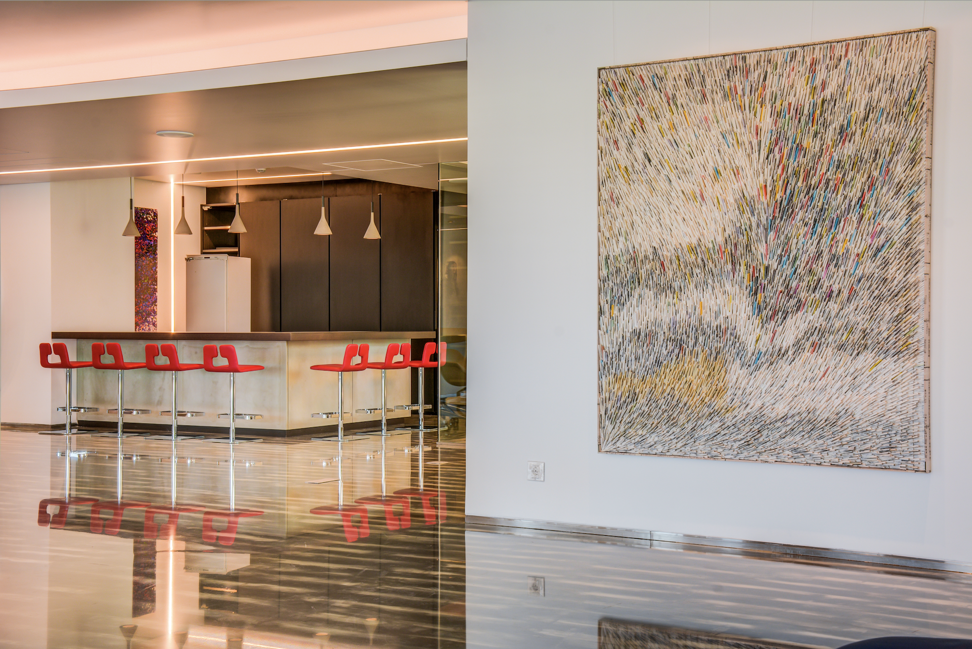 Modern indoor lounge area with a bar counter, red bar stools, contemporary pendant lighting, and an abstract textured artwork on the wall.