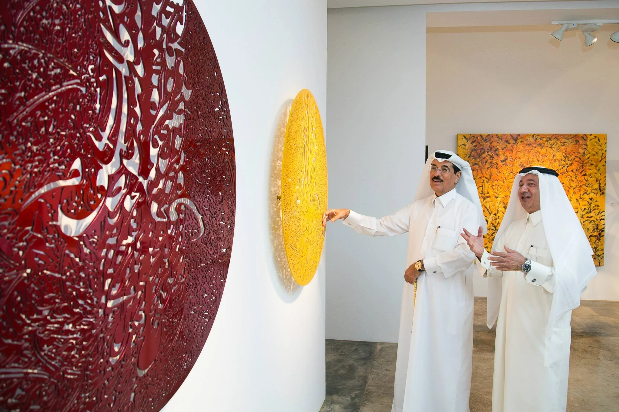 Two men in traditional Middle Eastern attire are viewing colorful artwork in an art gallery. One man is touching a yellow circular piece, while the other is gesturing with his hands, standing in front of various art pieces including a red circular ar